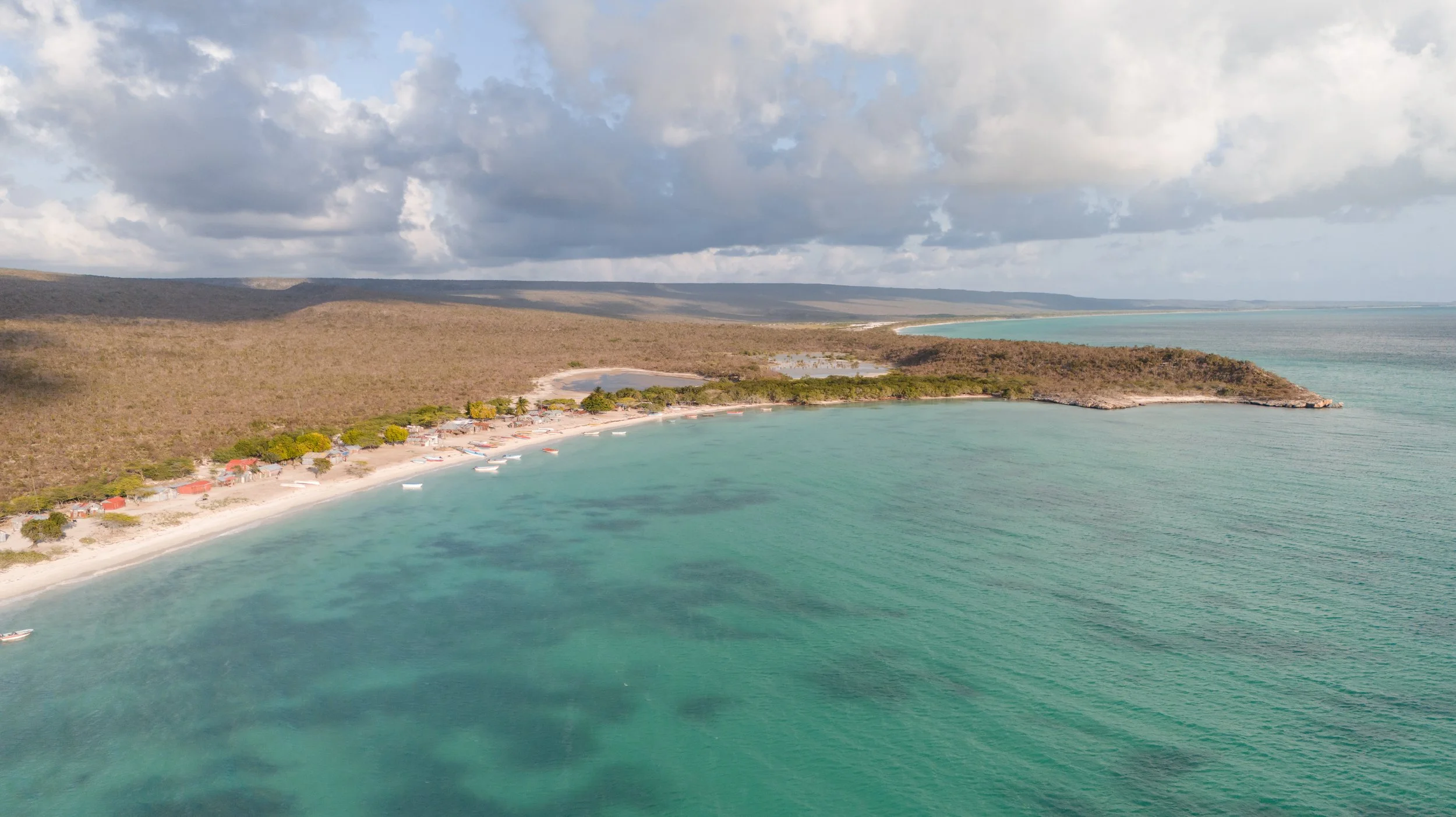 Vista aérea de una playa tropical con agua azul turquesa, arena blanca y pequeñas embarcaciones a lo largo de la costa. Al fondo, un paisaje árido y vegetación escasa bajo un cielo nublado.