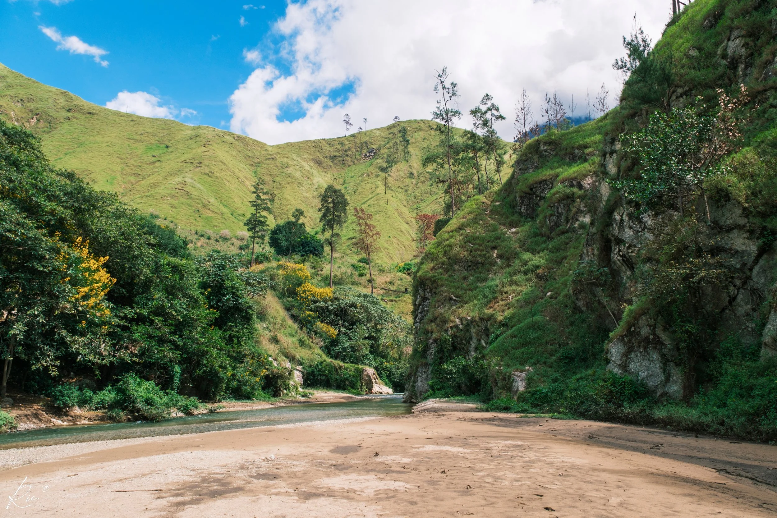 Paisaje de montaña con colinas verdes, cielo azul, nubes blancas y un río pequeño que fluye en el valle rodeado de vegetación.