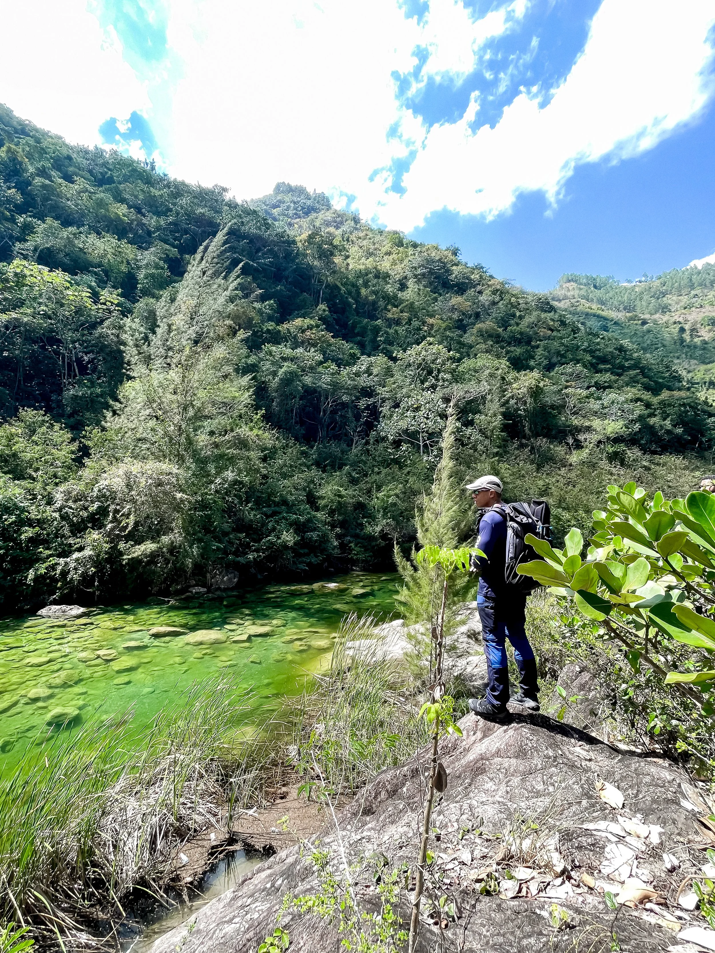 Persona de pie sobre una roca junto a un río de aguas verdes, rodeado de vegetación densa y montañas bajo un cielo azul con nubes.