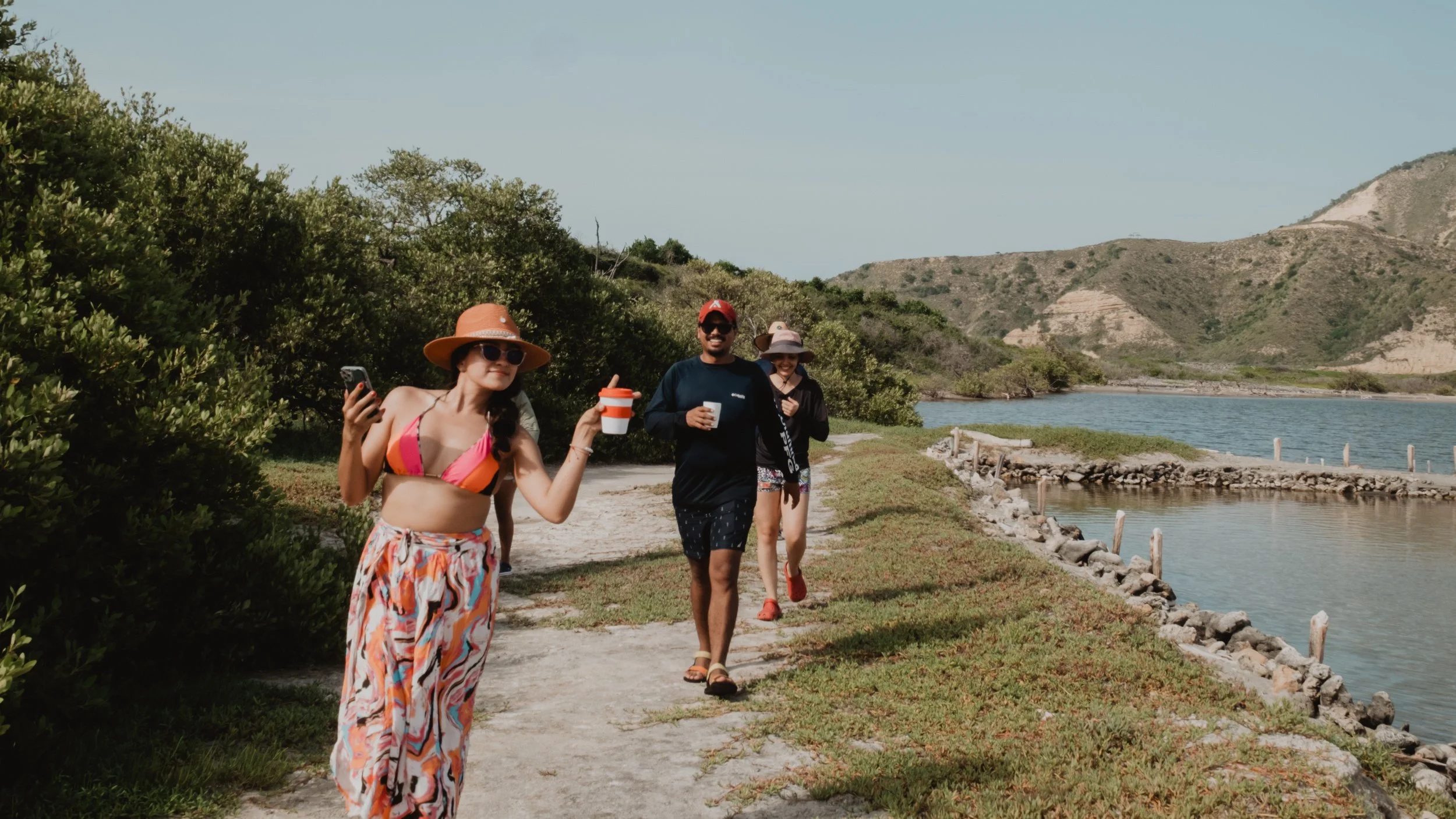 Tres personas caminando junto a un río en un entorno natural, con colinas al fondo. Una mujer lleva sombrero y ropa colorida, levantando una bebida. Otras dos personas también llevan sombreros y están sonriendo. Hay vegetación a su alrededor y un cam