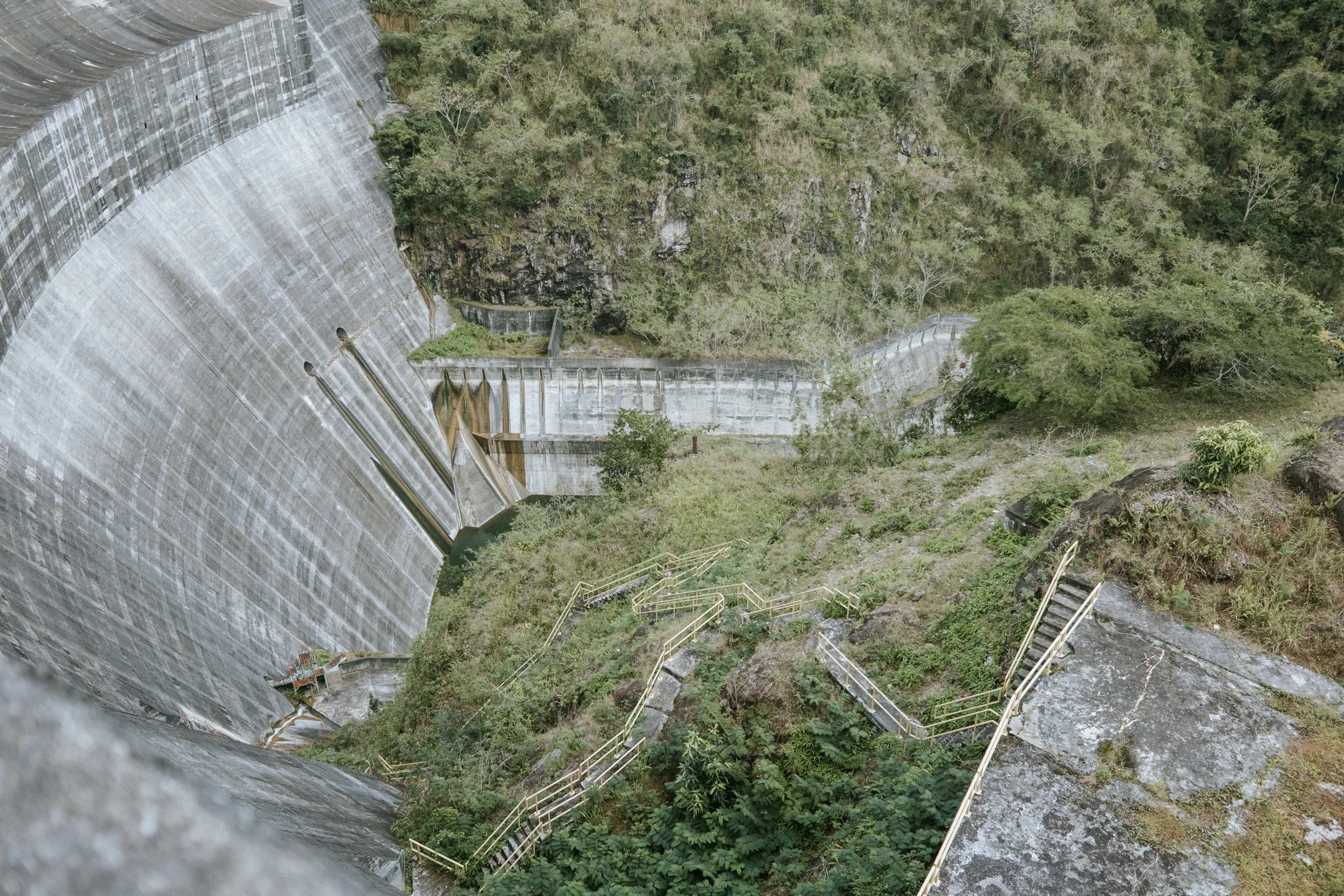 Presa de concreto en un paisaje verde y montañoso, con escaleras y barandillas visibles.