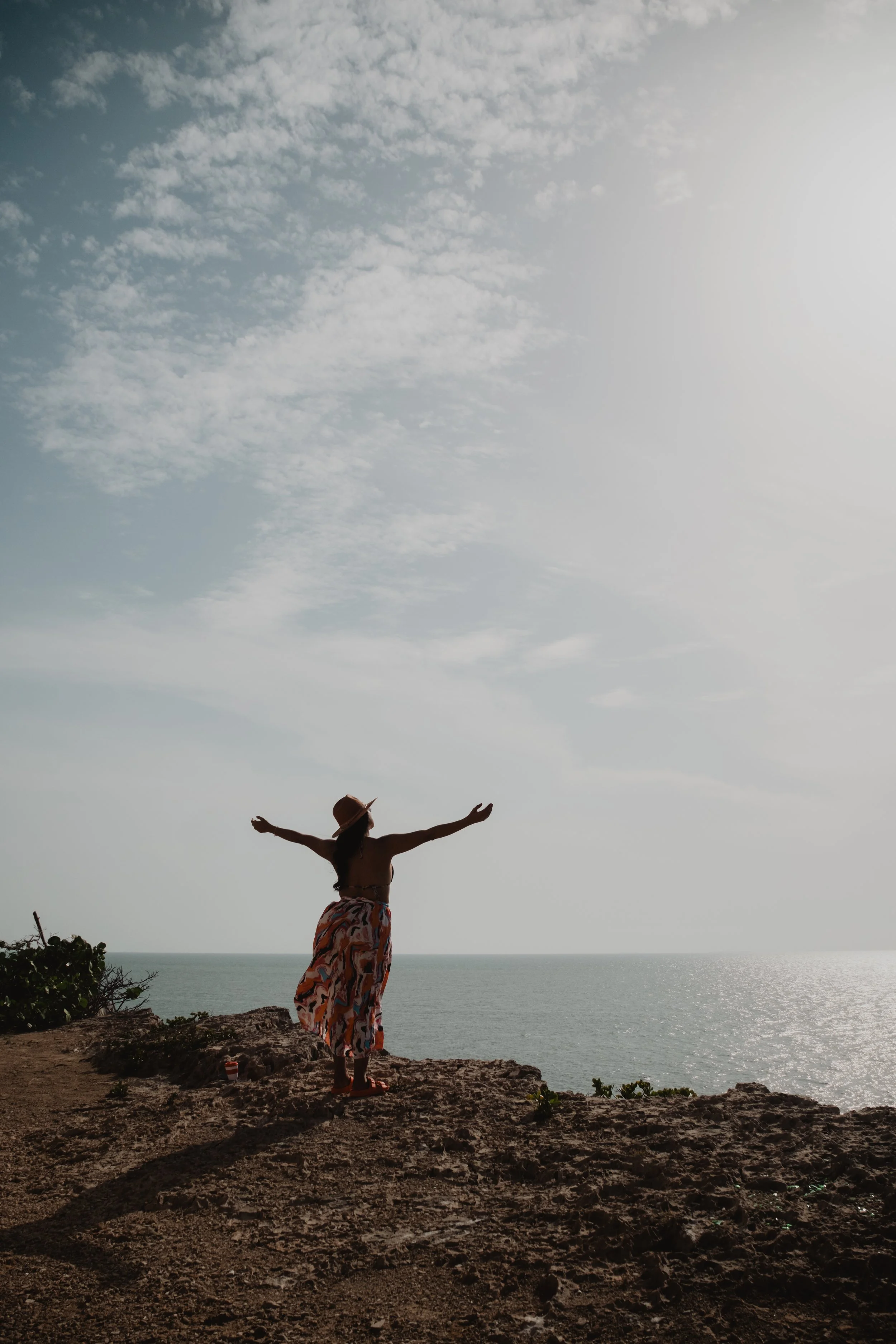Mujer de pie con los brazos abiertos en un acantilado junto al mar, vistiendo un sombrero y una falda colorida, bajo un cielo parcialmente nublado.