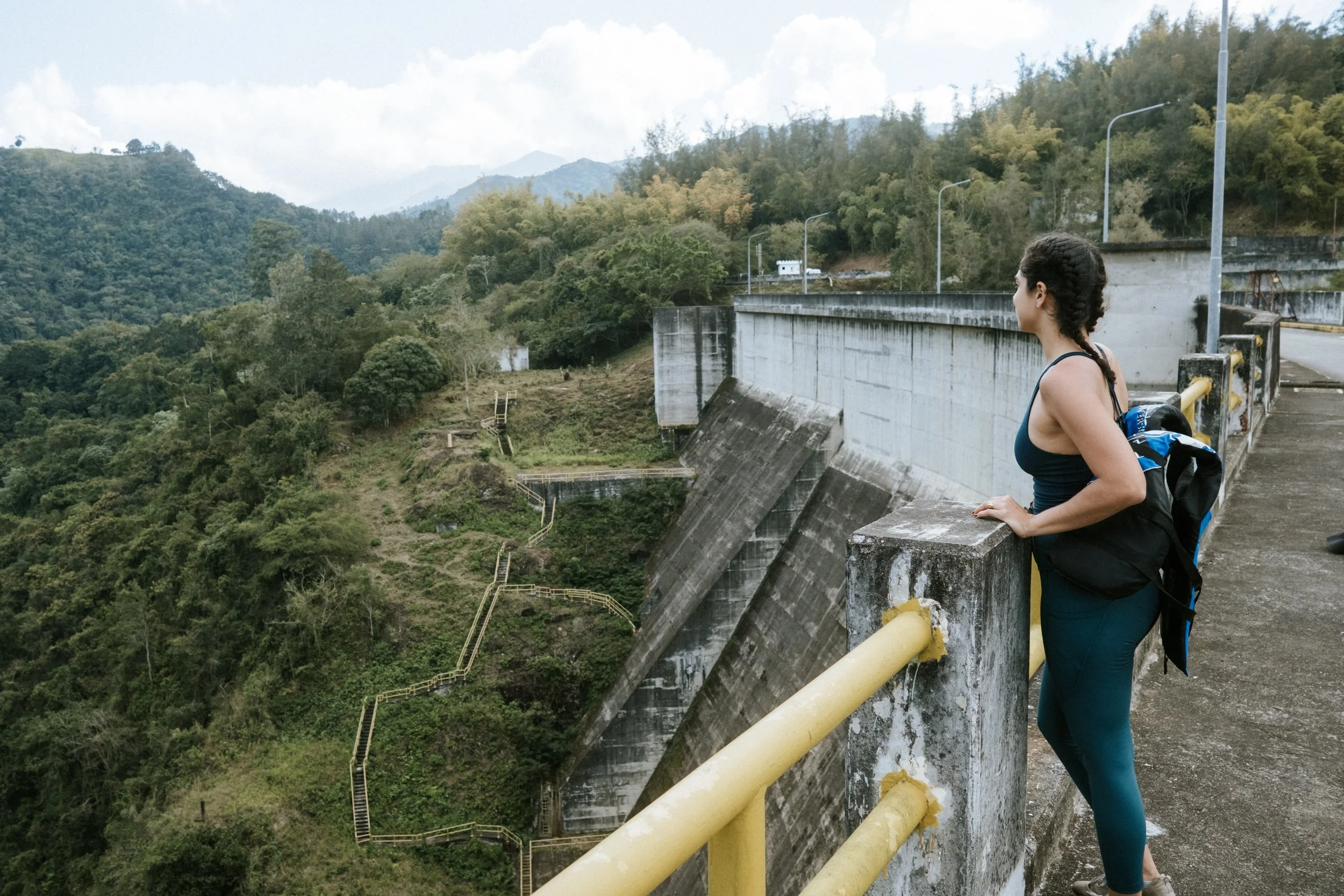 Una persona observando un paisaje montañoso junto a una estructura de hormigón, posiblemente una presa. La vegetación es abundante y hay senderos visibles en el terreno. El cielo está parcialmente nublado.