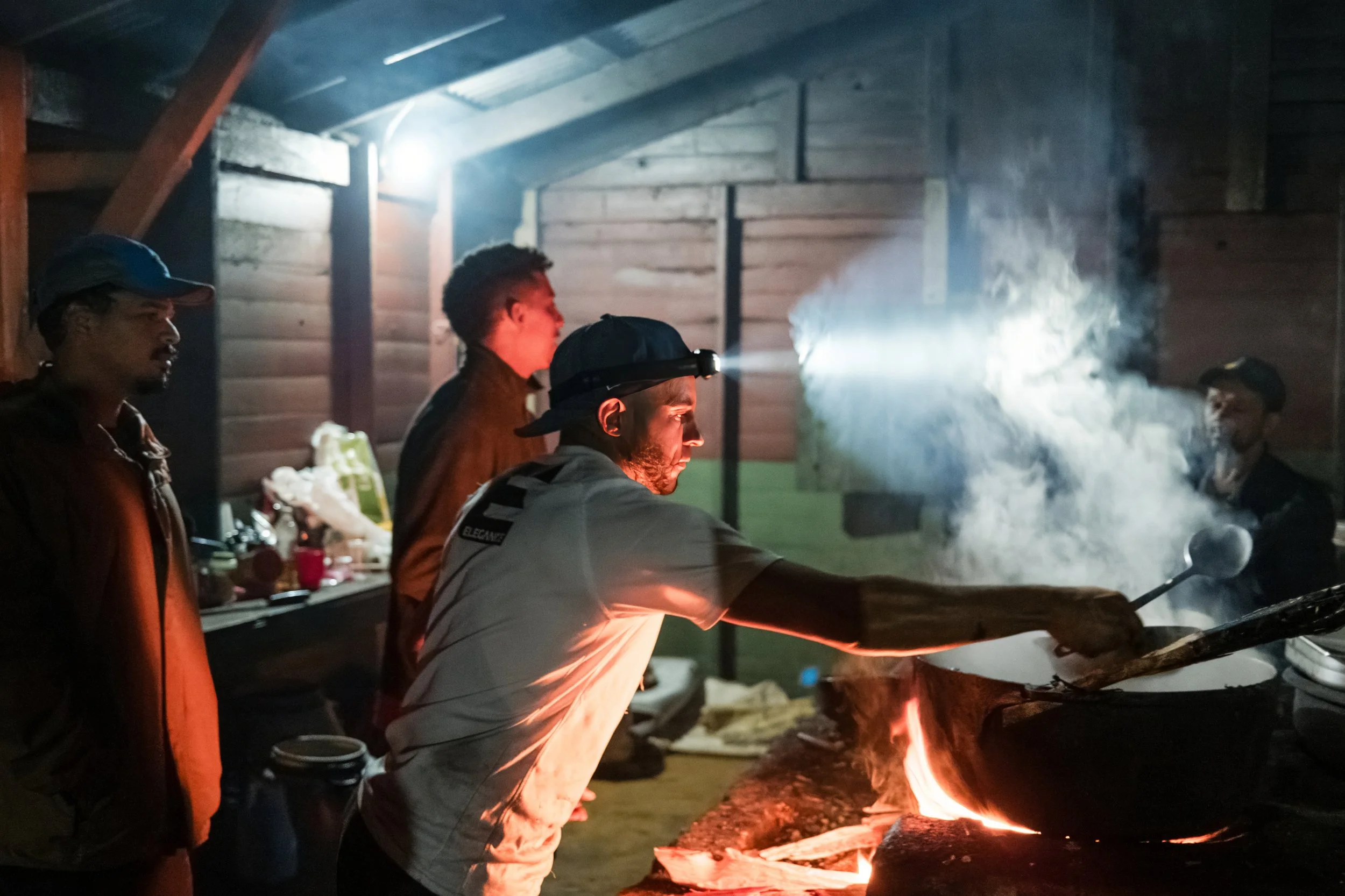 Grupo de personas cocinando en una cocina rústica con fogón y utensilios de cocina, en un ambiente con poca luz.