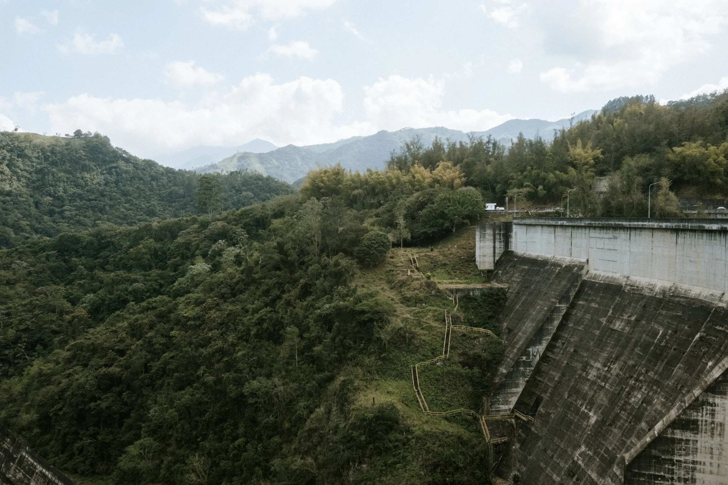 Vista panorámica de una presa de concreto con montañas y vegetación abundante en el fondo, bajo un cielo parcialmente nublado.
