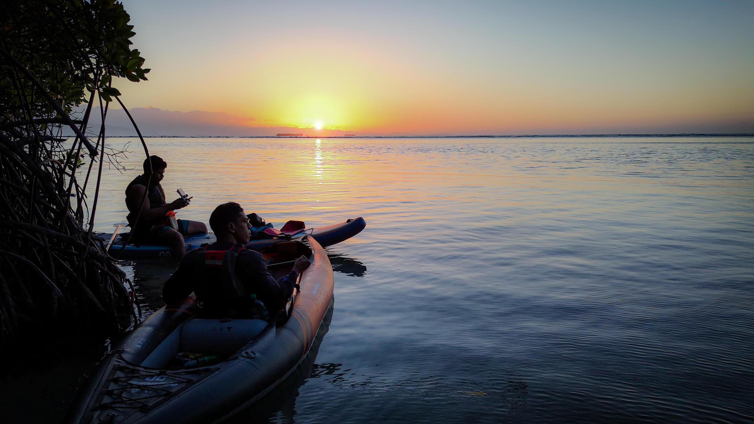 Dos personas en kayaks disfrutando de una puesta de sol en el agua, con árboles y un cielo claro en el fondo.