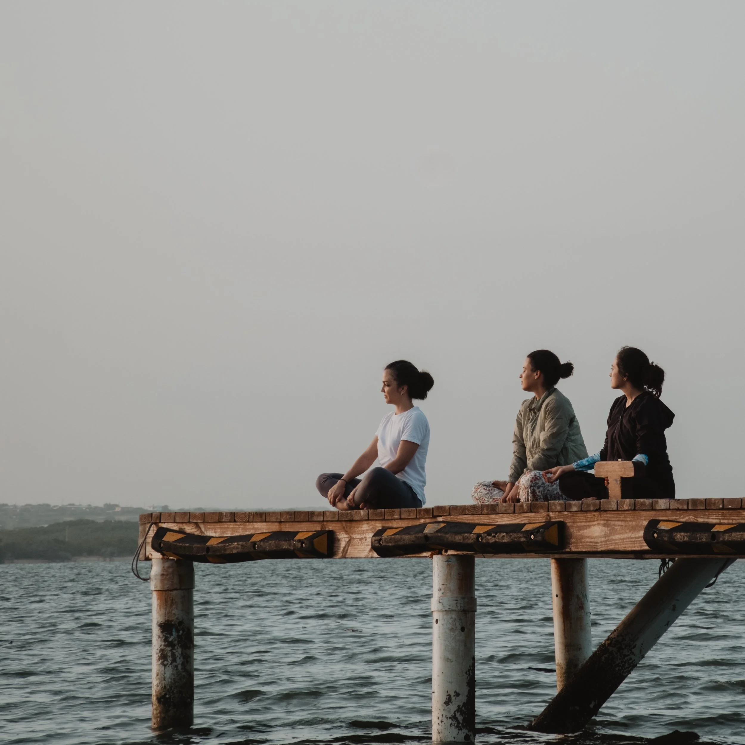 Tres personas sentadas sobre un muelle de madera mirando el horizonte sobre el agua.