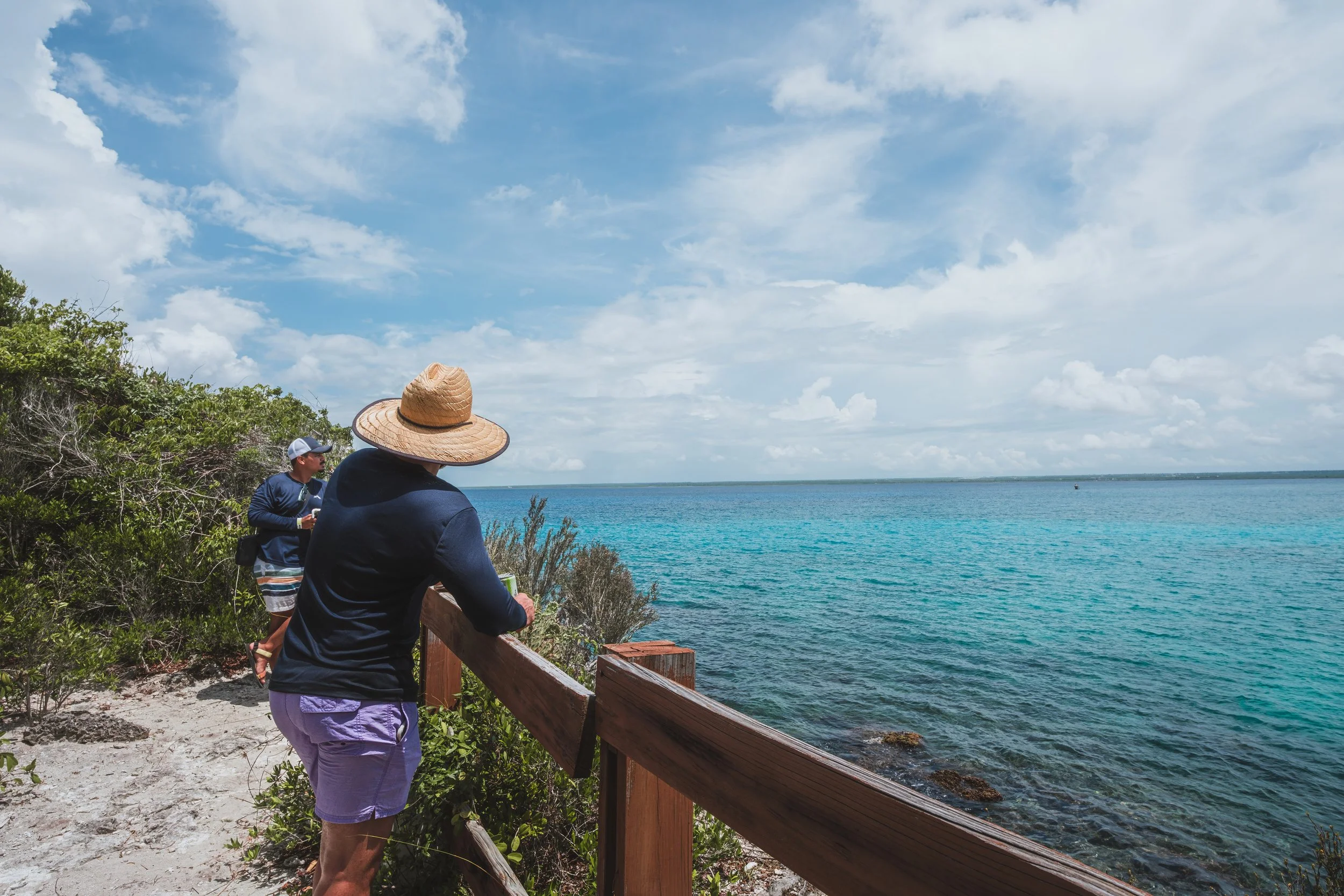 Dos personas observando el mar desde un mirador con baranda de madera, vegetación alrededor, cielo azul con nubes.