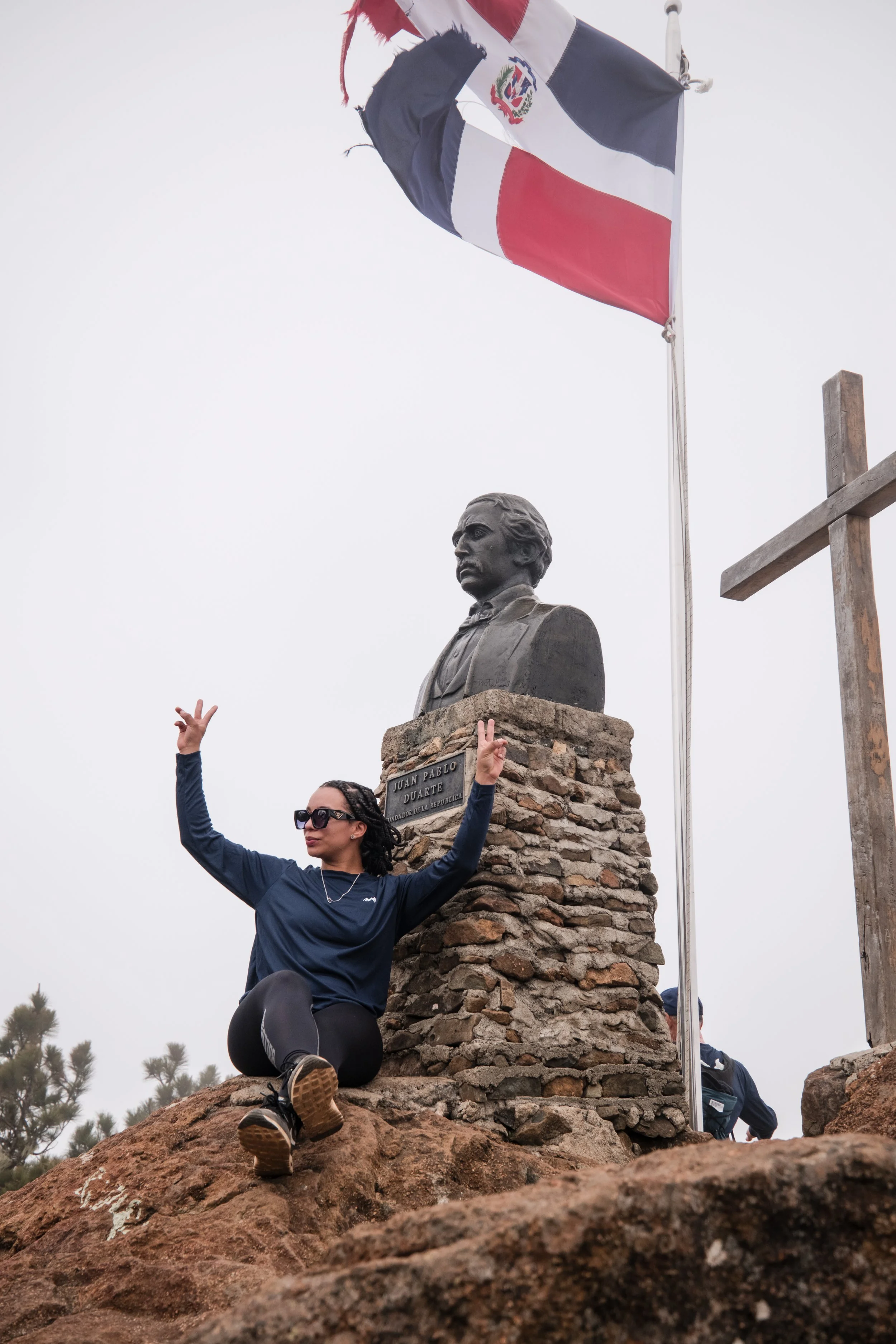 Persona sentada al lado de un busto de Juan Pablo Duarte, con la bandera dominicana ondeando arriba y una cruz de madera al fondo.