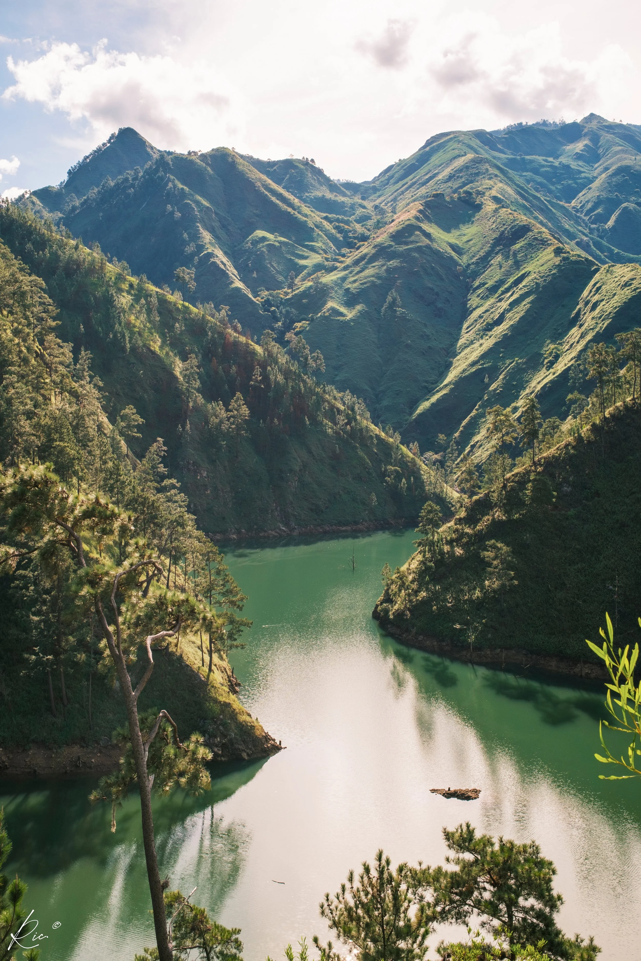 Paisaje montañoso con colinas verdes y un lago rodeado de árboles. El cielo está parcialmente nublado con luz solar suave.