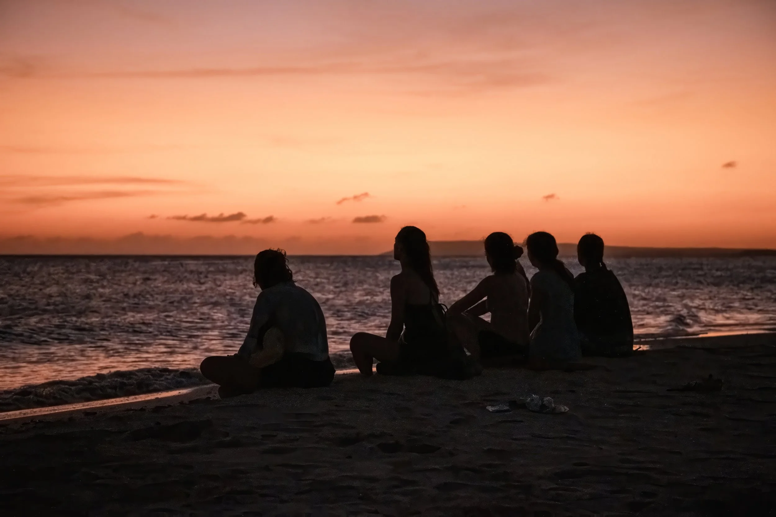 Grupo de personas sentadas en la playa durante el atardecer, mirando el mar.