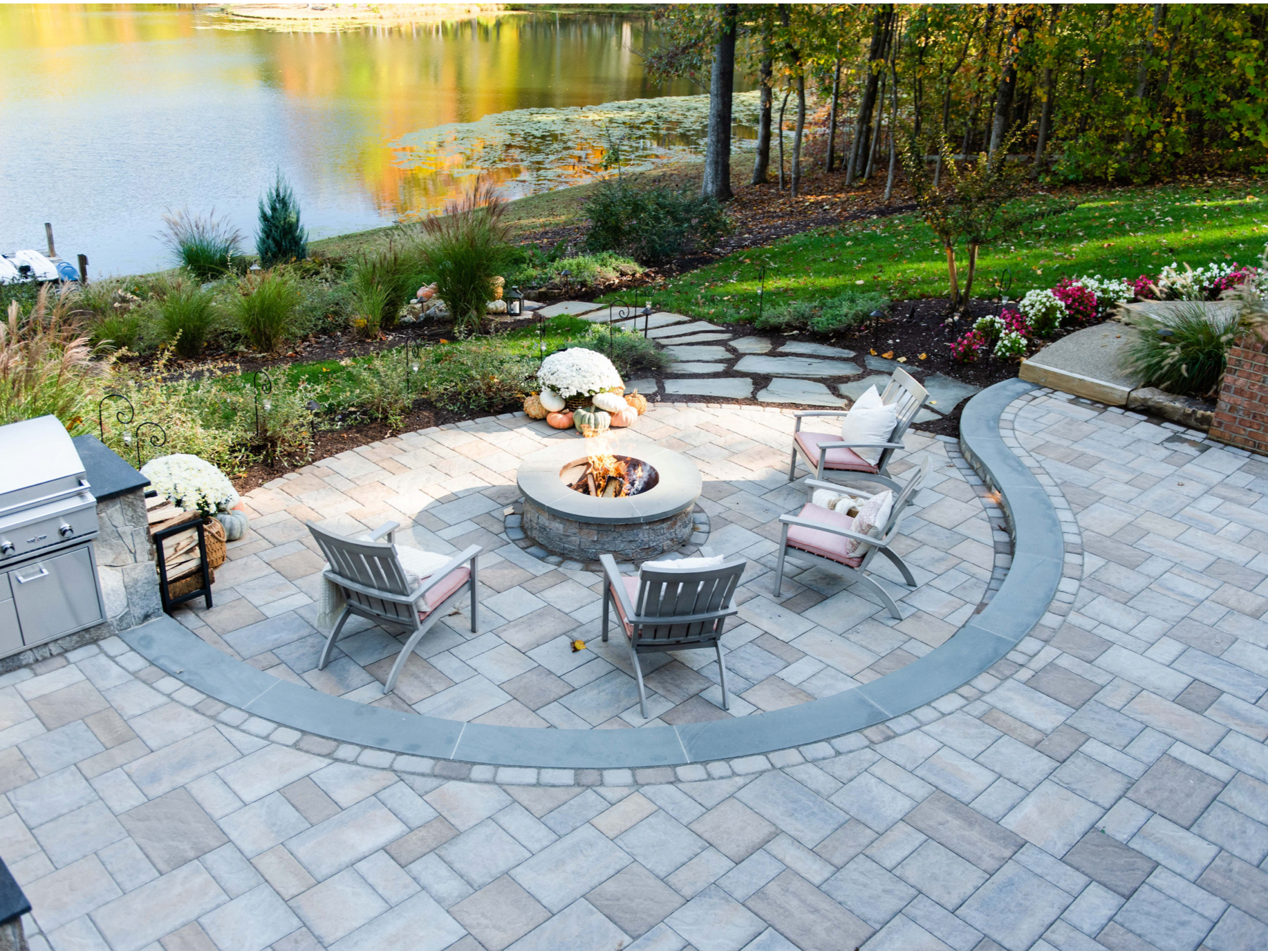 A backyard patio overlooks a pond with yellow and green foliage. The patio has a circular fire pit with four chairs arranged around it, decorated with pillows. There are fall-themed decorations like pumpkins and flowers on the patio and garden plants in the background.