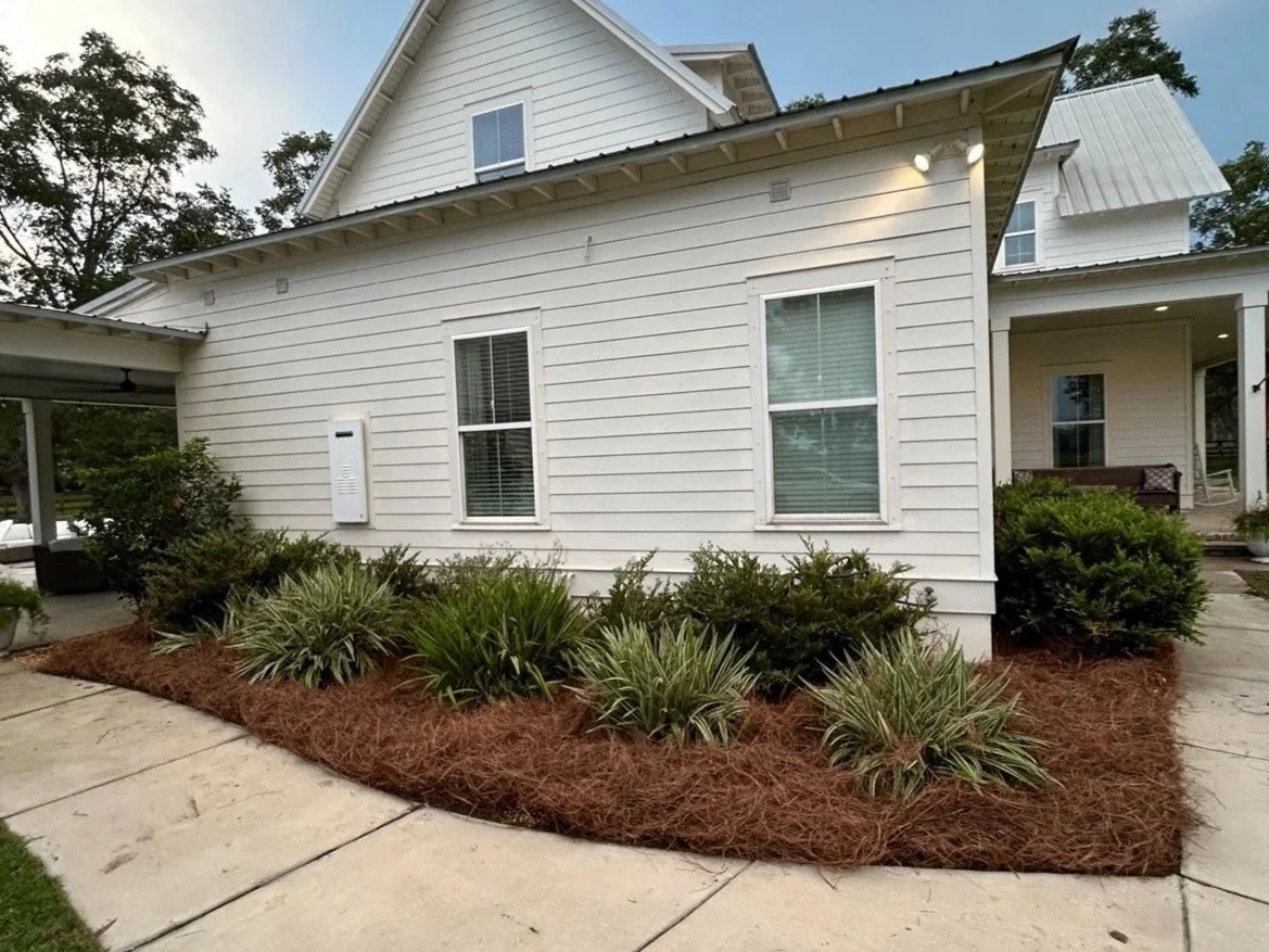 White house with garden and shrubs in the front yard, sidewalk leading up to the house, and a porch with seating area