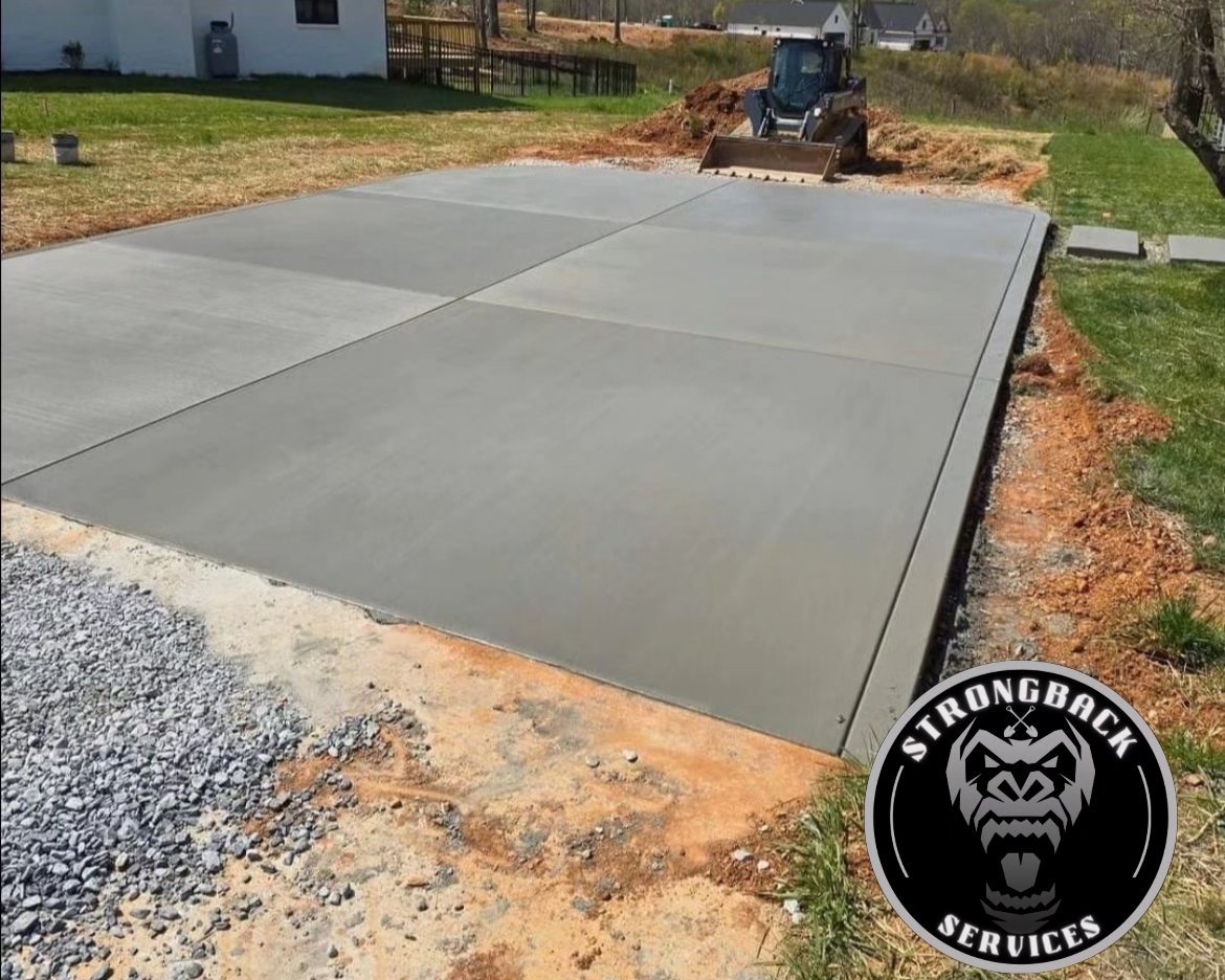 Newly poured concrete slab in a backyard with a bulldozer nearby, surrounded by grass and dirt, with a house and trees in the background, and a Strongback Services logo in the bottom right corner.