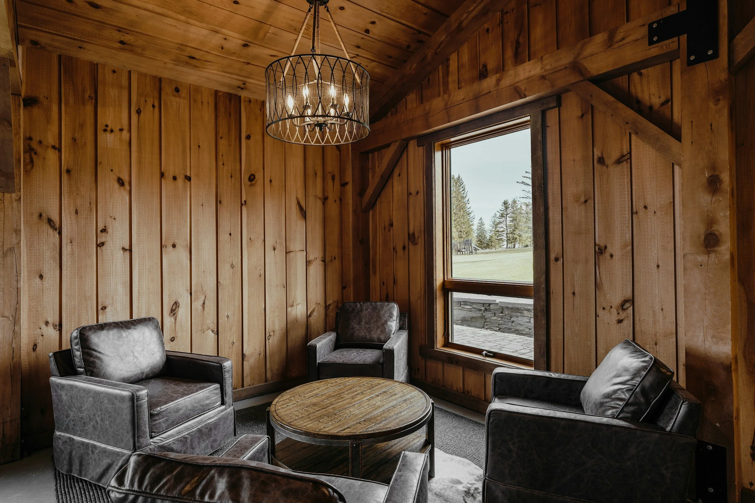 Inside a rustic wooden room with a large window showing a view of trees outside, a chandelier hanging from the ceiling, and black leather chairs around a round wooden table.