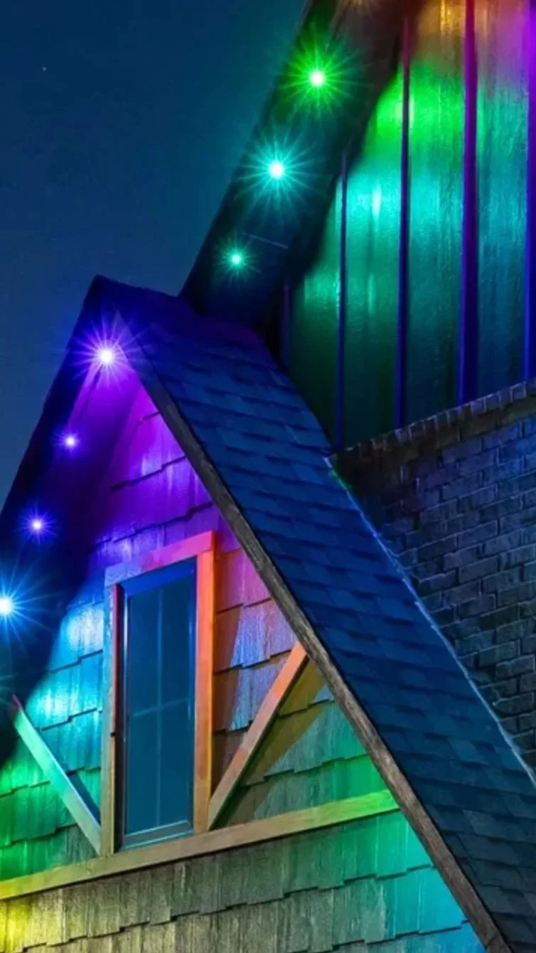 Nighttime view of a house with colorful rainbow lights illuminating the exterior, including shingles, window, and roof.