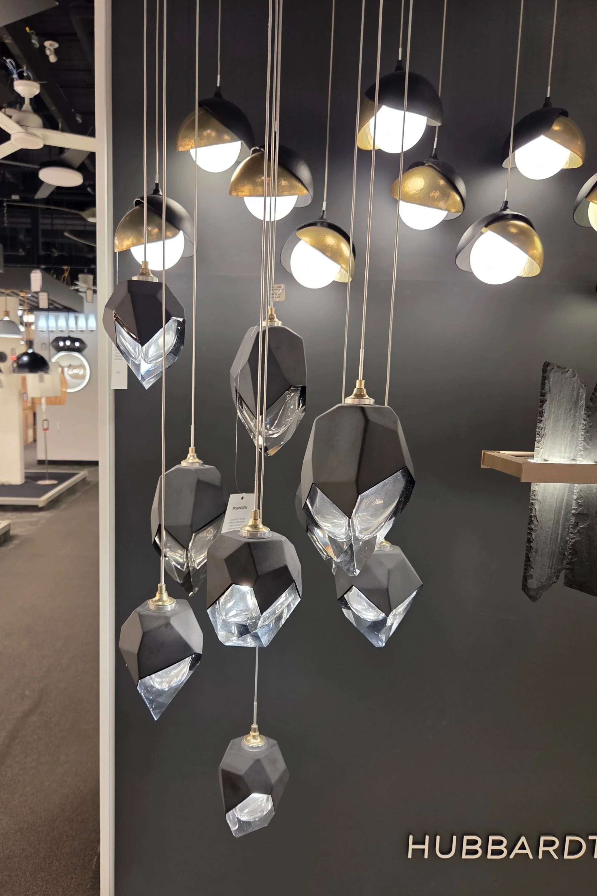Multiple modern hanging pendant lights with black and gold accents and geometric crystal elements are displayed against a dark wall in a lighting store.