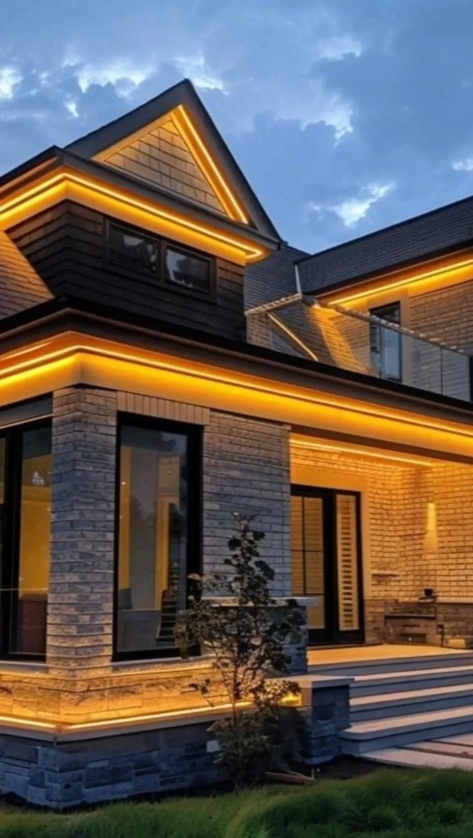 A modern house with exterior brick walls, illuminated with warm yellow lighting along the roof edges and around the windows, with a small tree in front and a staircase leading to the entrance, captured during dusk.