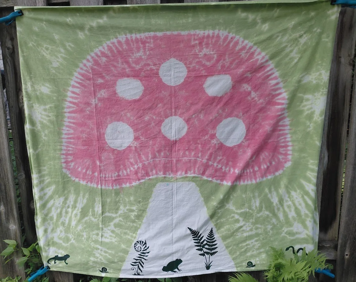 Tie-dye fabric with a pink mushroom and white spots, set against a green background with black silhouettes of ferns and insects at the bottom.