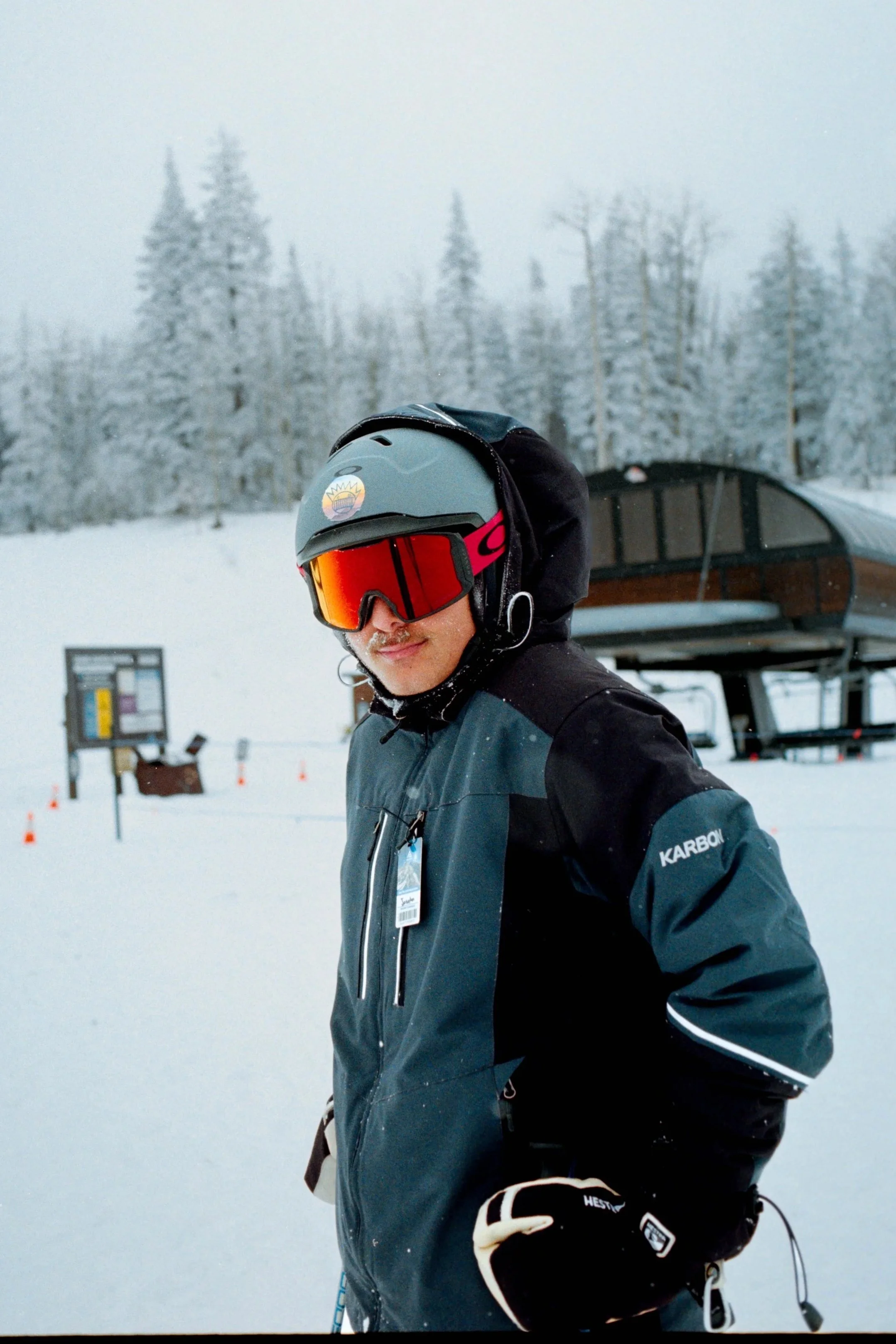 Person wearing ski gear with a helmet and goggles on a snowy mountain with trees and a ski lift in the background.