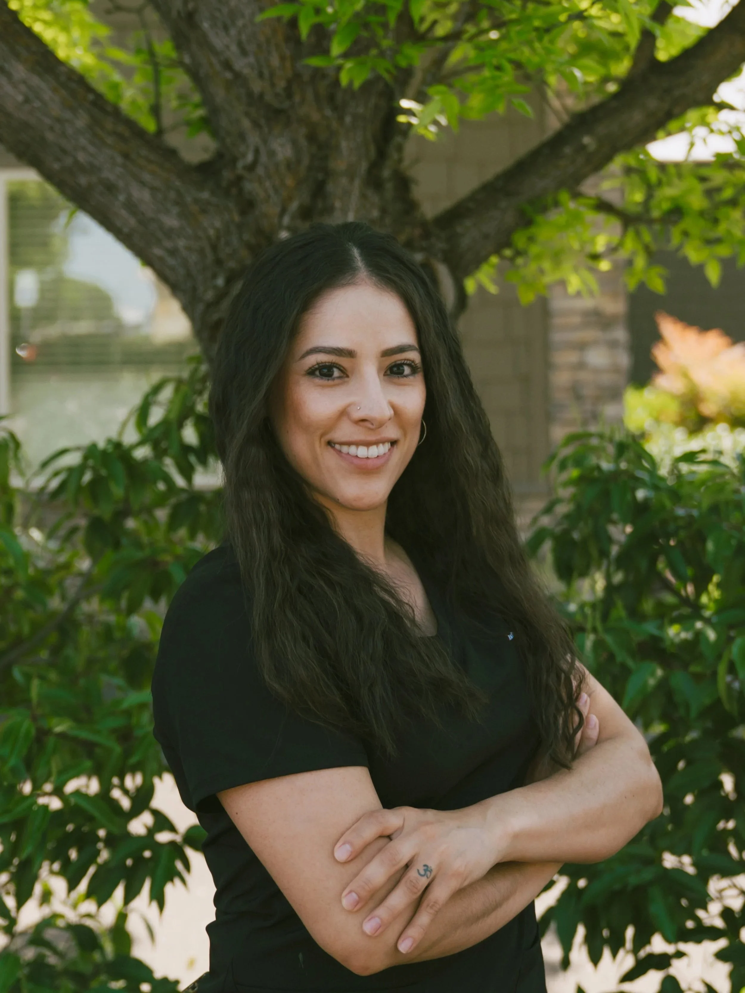 A woman with long dark wavy hair, wearing a black shirt, standing outdoors in front of a tree with green leaves, smiling with arms crossed.