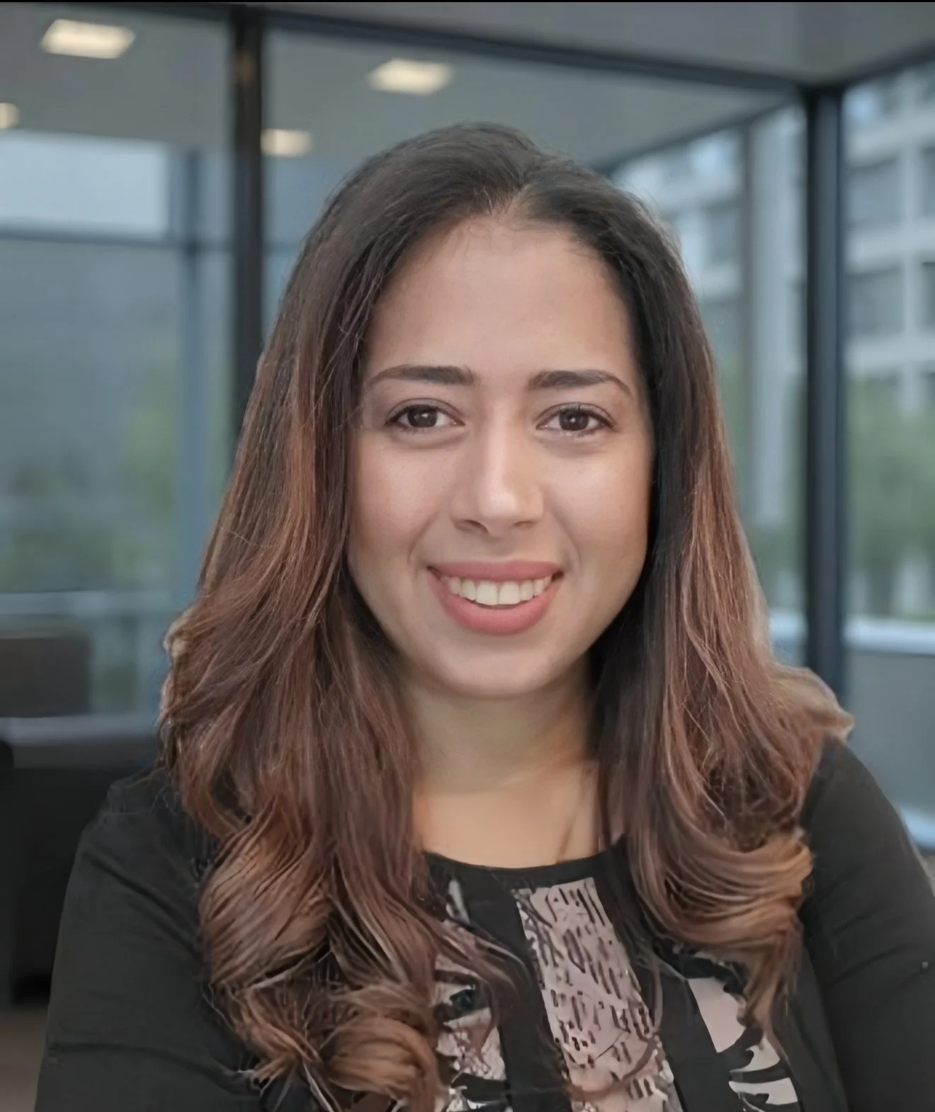 A smiling woman with long, wavy brown hair with lighter tips, wearing a patterned top and a black blazer, sitting in an office with large windows.