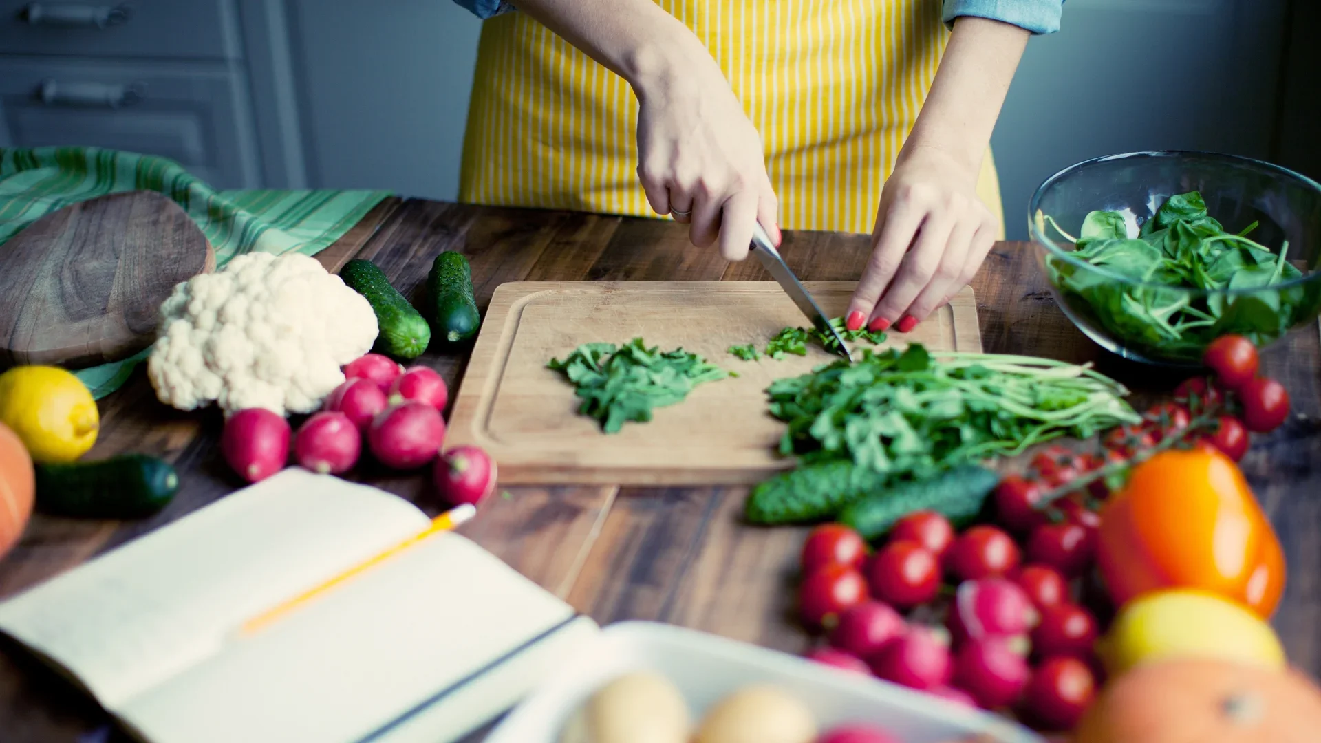 Person chopping leafy greens on a wooden cutting board, surrounded by various fresh vegetables including radishes, cauliflower, cucumbers, and bell peppers, on a kitchen counter.