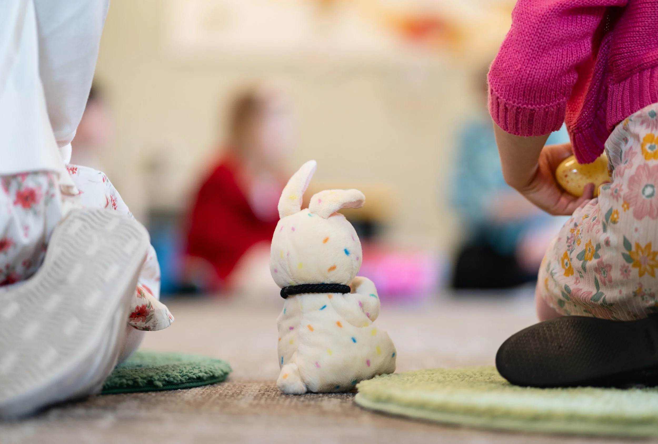 Close-up of a small stuffed bunny toy with rainbow polka dots, placed on a carpeted floor between two children, one on each side, in a room with a blurred background.