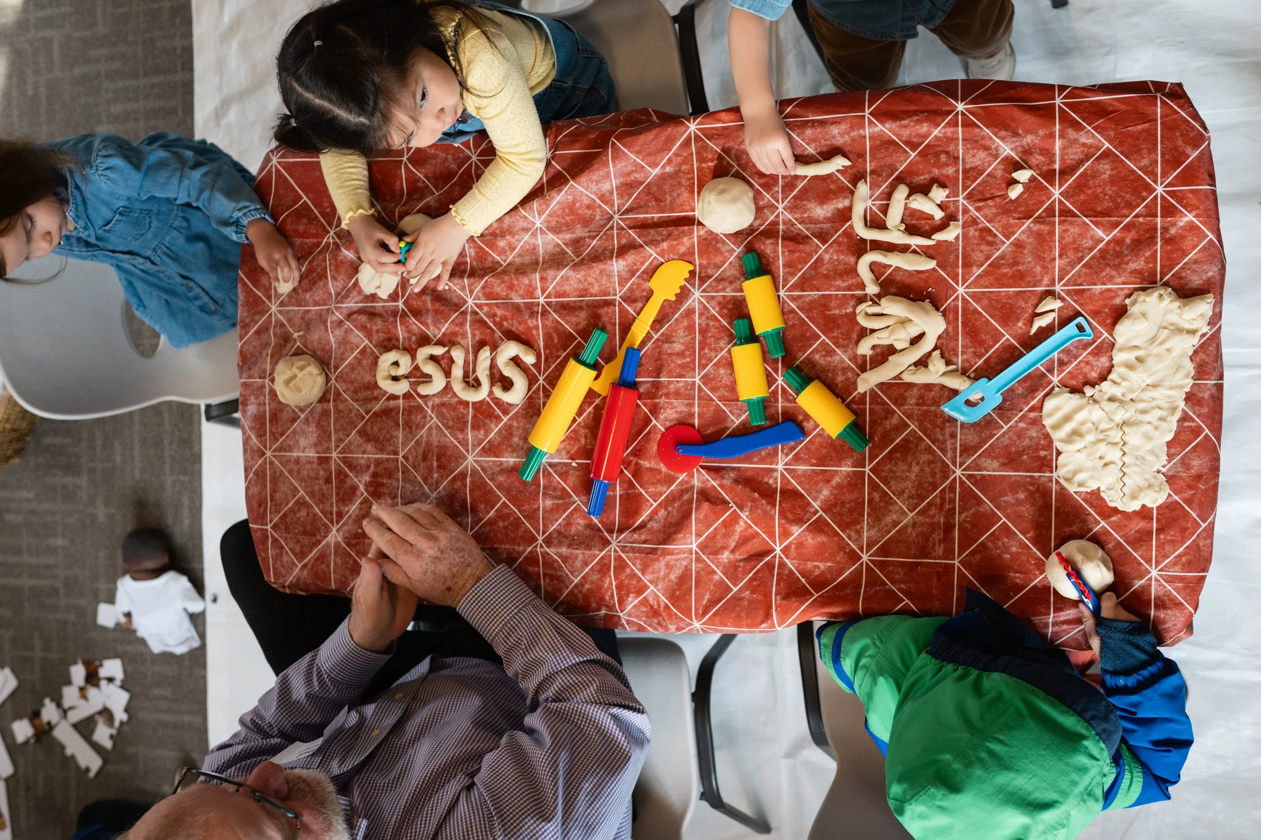 Children and an adult creating sculptures with white modeling clay on a red tablecloth, using various modeling tools.