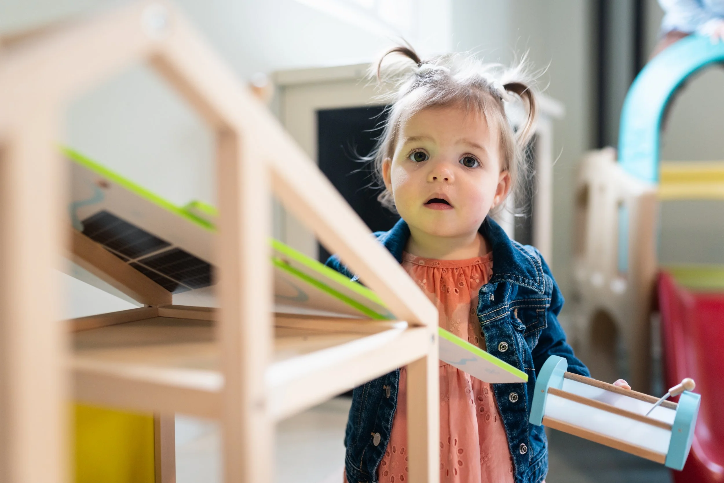 A young girl with pigtails and a denim jacket standing behind a wooden toy.