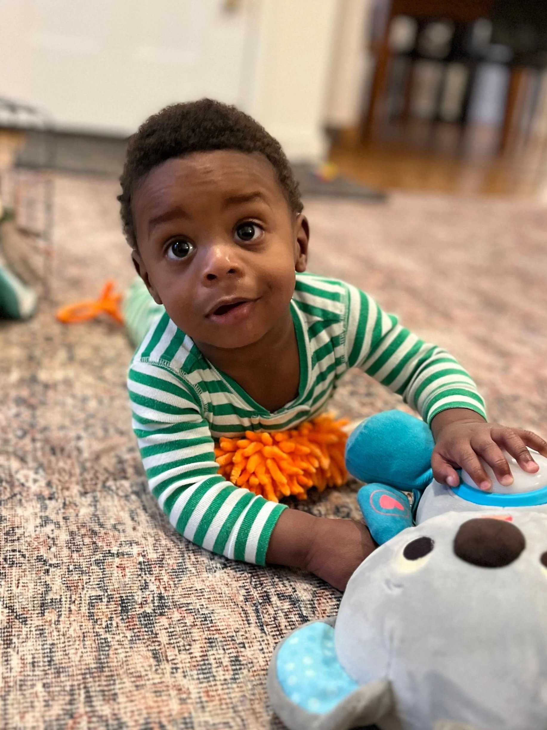 A young child with dark hair, wearing a green and white striped shirt, is lying on a carpeted floor, engaging with plush toys and a blue ball, with a background of a bookshelf and a door.