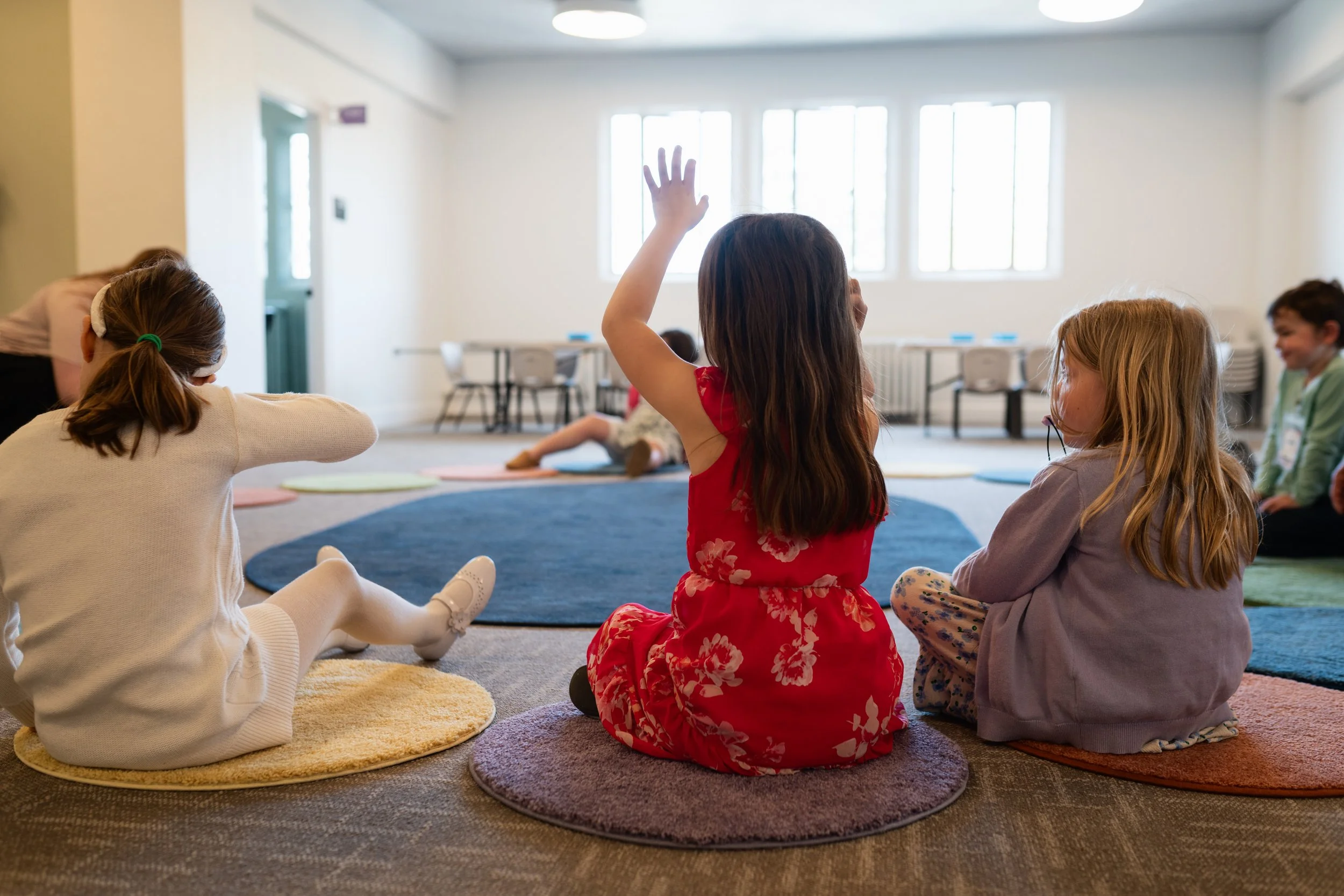 Children sitting on colorful circular rugs in a classroom, with one girl raising her hand and others listening.