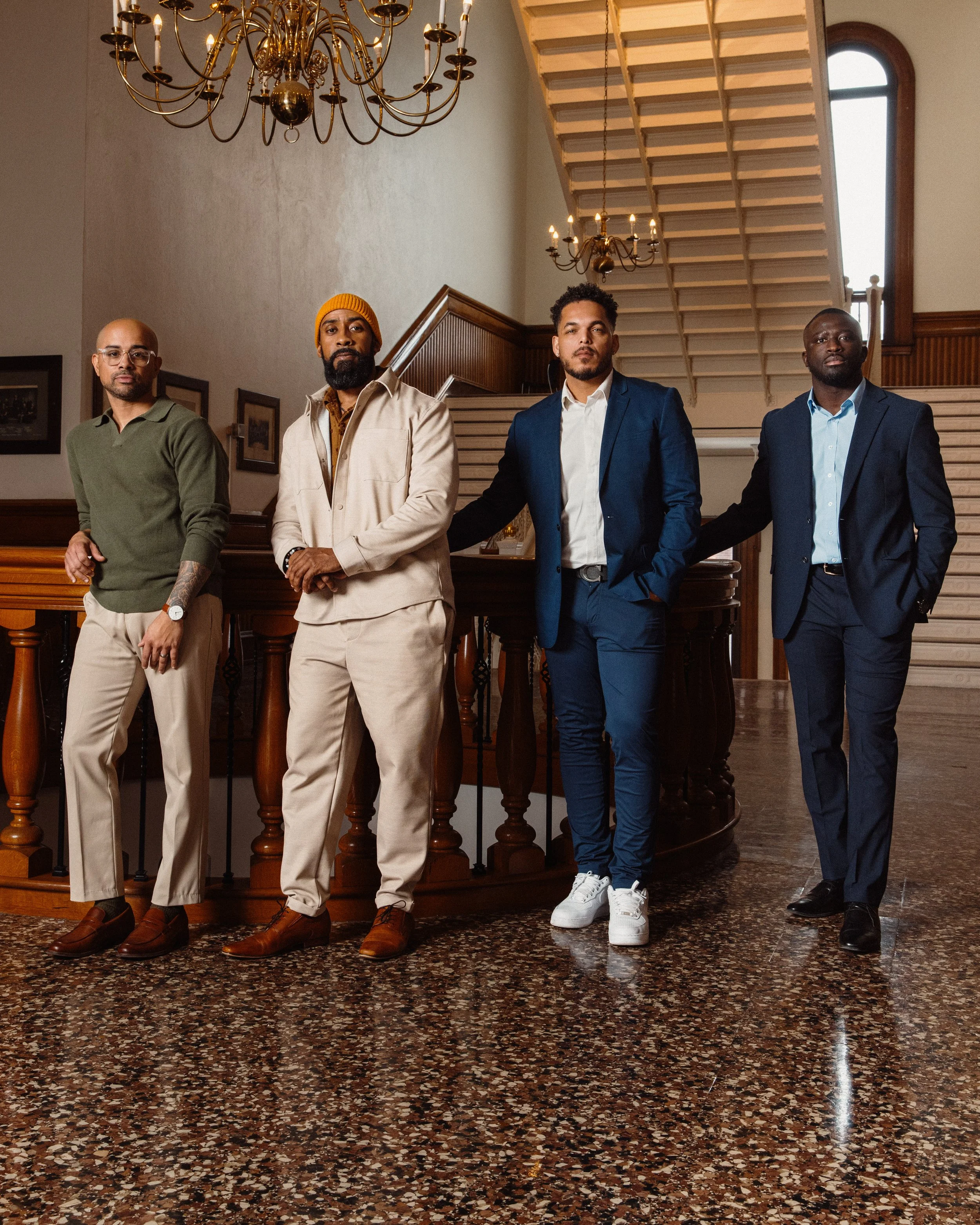 Four men standing inside a building with a staircase, chandelier, and framed pictures on the wall behind them. They are dressed in smart-casual to formal attire. Nelson Fernandes, Rui Lopes, Salo Afonseca and Francois Diatta