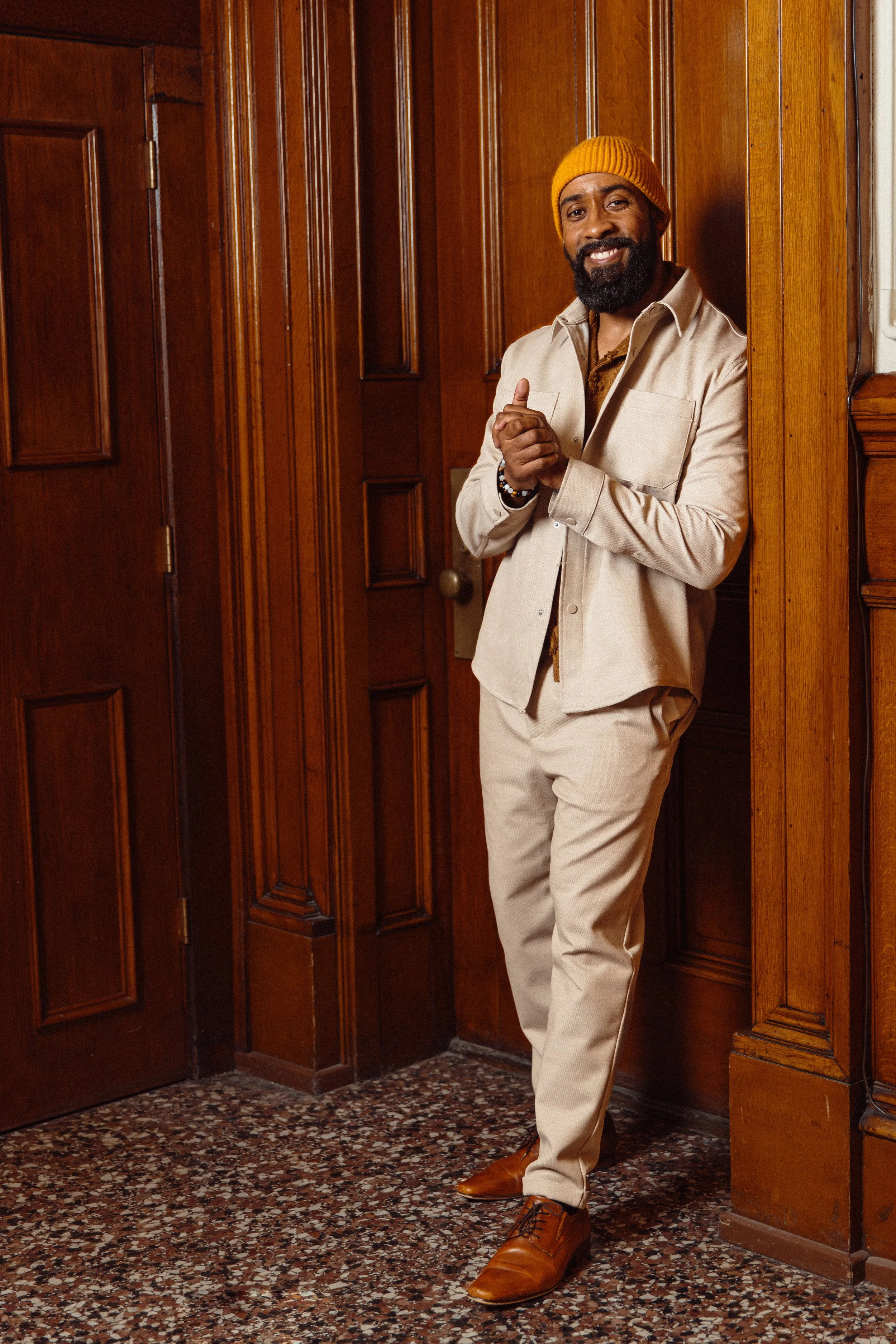 Rui Lopes with a beard and yellow beanie, wearing a beige suit, standing in a wooden-paneled room, smiling, with one hand raised and the other holding his wrist.
