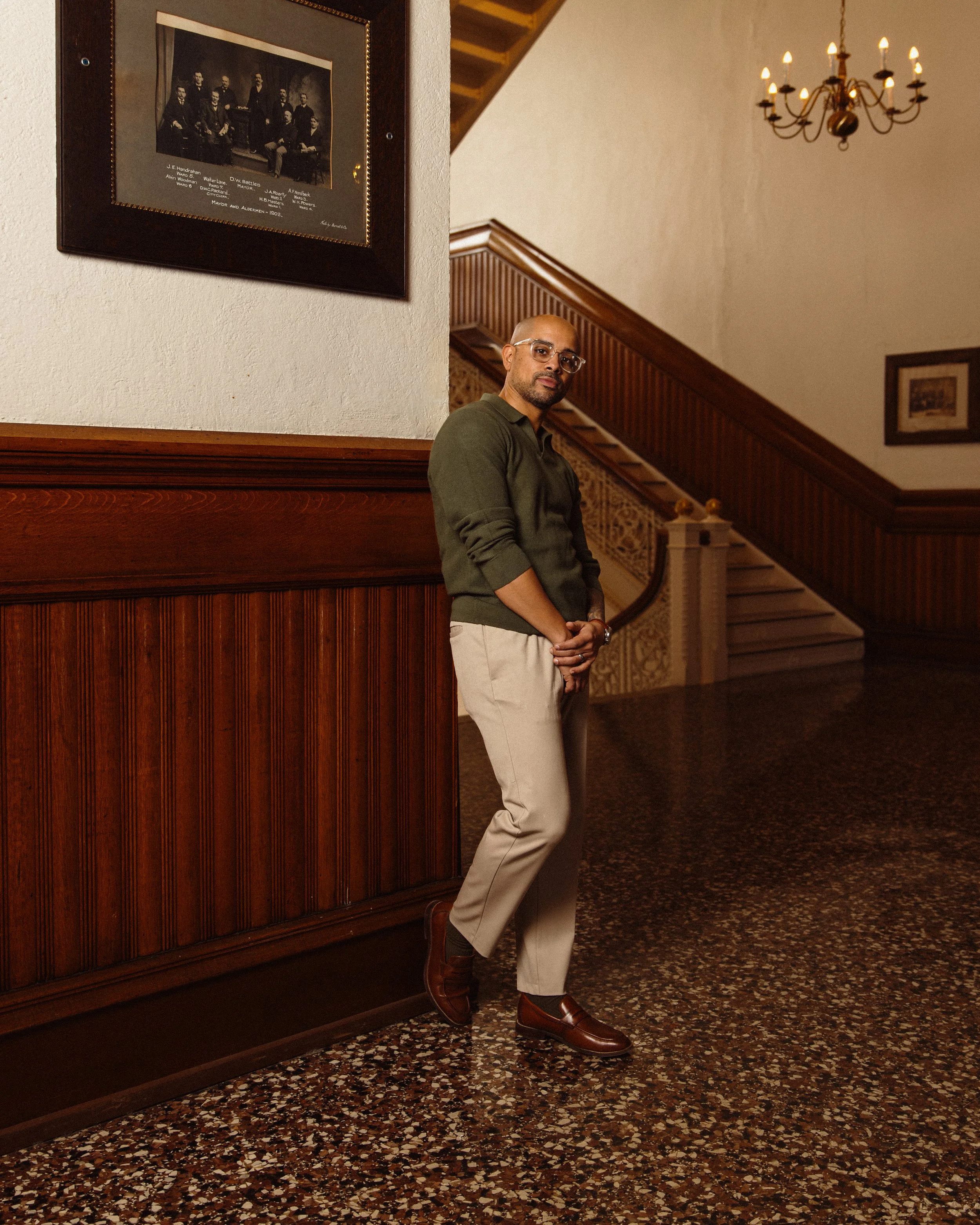 Nelson Fernandes, Deputy Chief of Staff with glasses, a bald head, wearing a green sweater, beige pants, and brown shoes, standing in a warmly lit indoor setting with wood paneling and a staircase in the background.