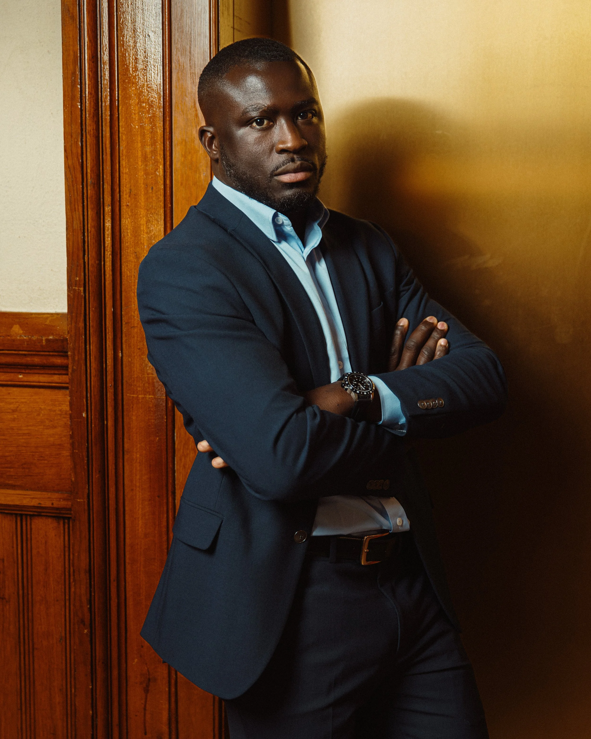 Francois Diatta in a business suit standing with his arms crossed, leaning against wooden wall panels.