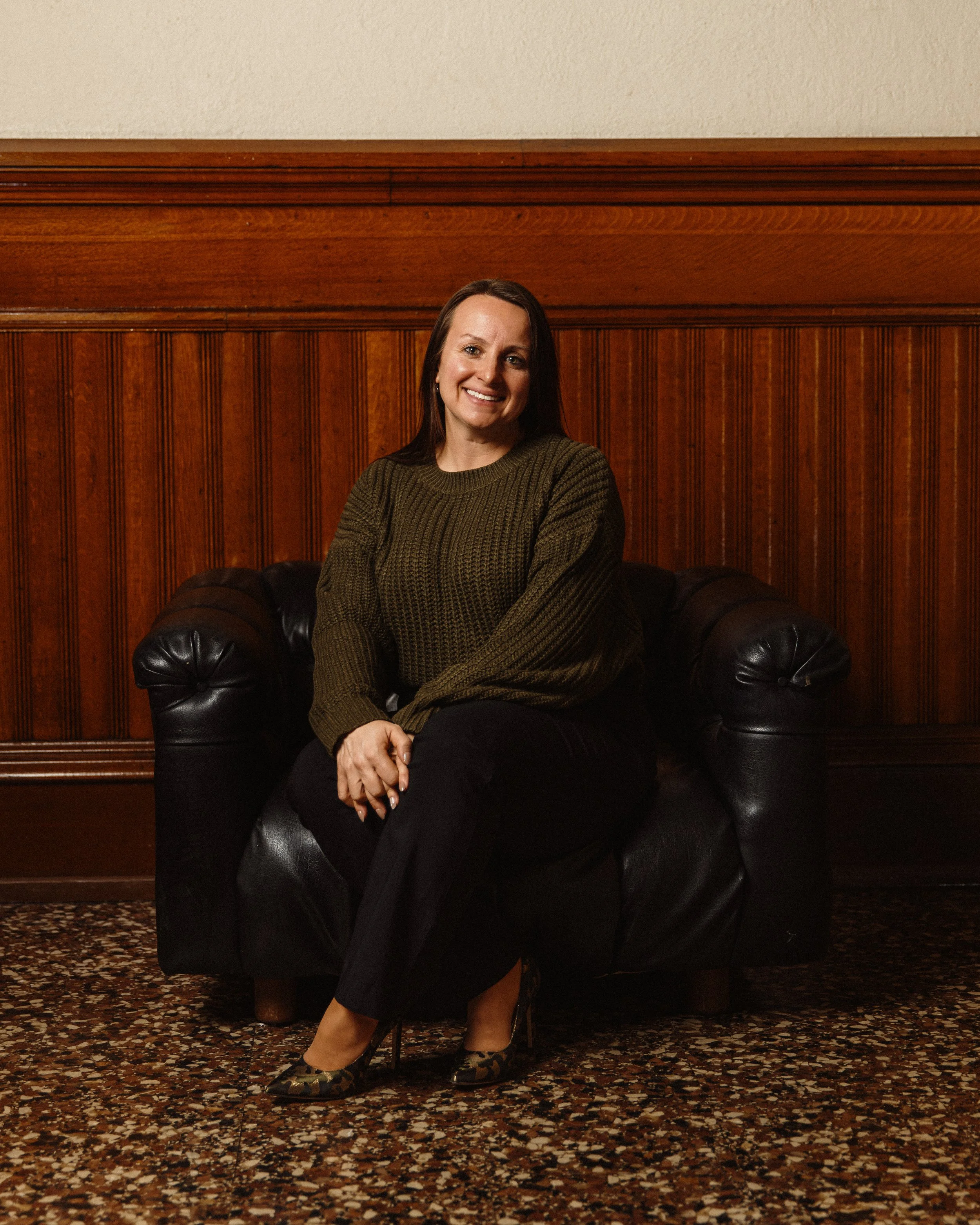 Katie Balboni with Brockton High dark hair smiling, sitting on a dark leather sofa, wearing a green sweater, black pants, and leopard print high heels, in front of a wooden paneled wall.
