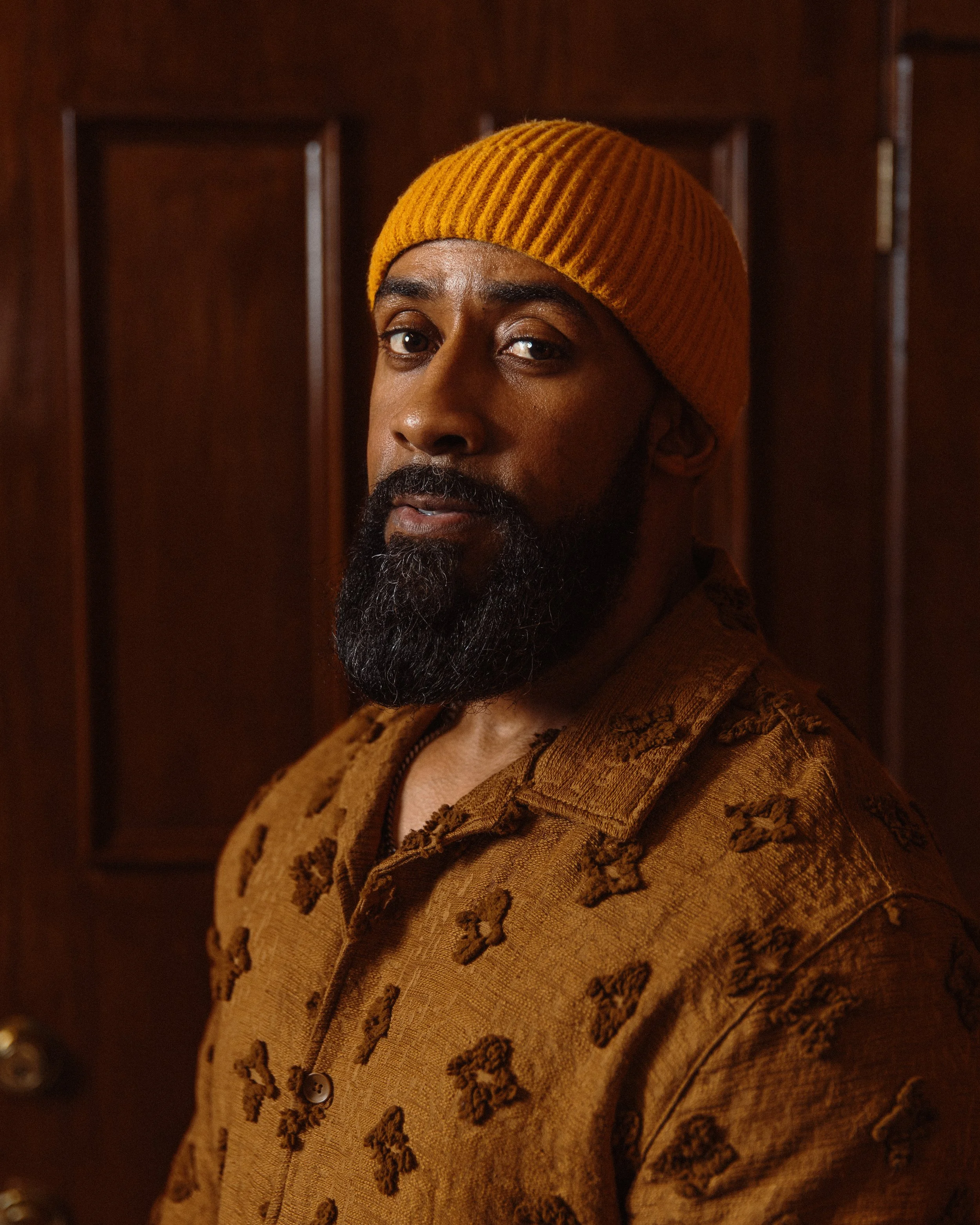 Rui Lopes of Anawan Studios with a dark beard and mustache wearing a brown jacket with embroidered and textured details and a mustard yellow knit beanie, standing in front of a dark wooden background.