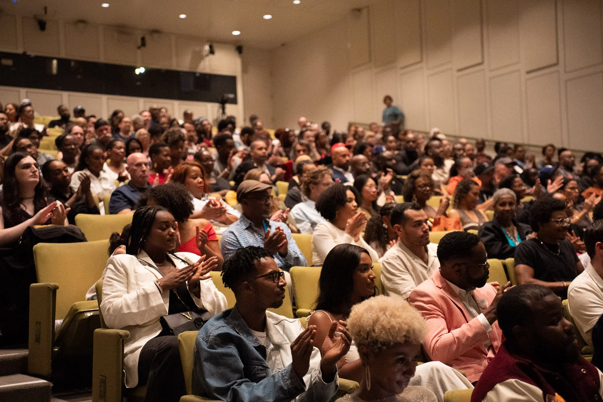 A large audience seated in a theater or auditorium, attentively listening and clapping, with diverse individuals including women and men of various ages and ethnicities.