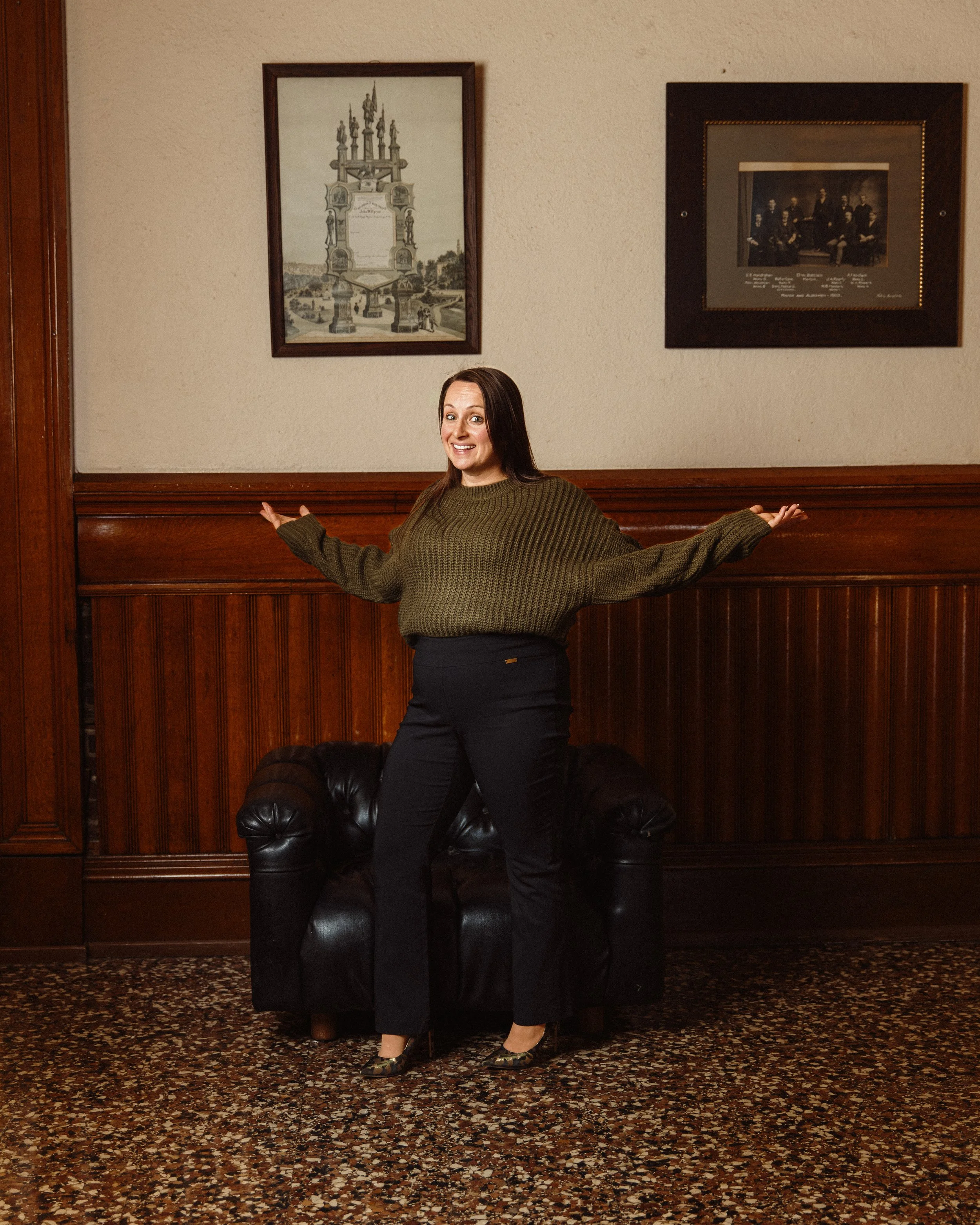Woman with brown hair smiling, standing in a room with wood-paneled walls, posing with her arms outstretched. She's wearing a green sweater and black pants, with black high heels. Behind her is a black leather sofa, and above her are two framed pictu