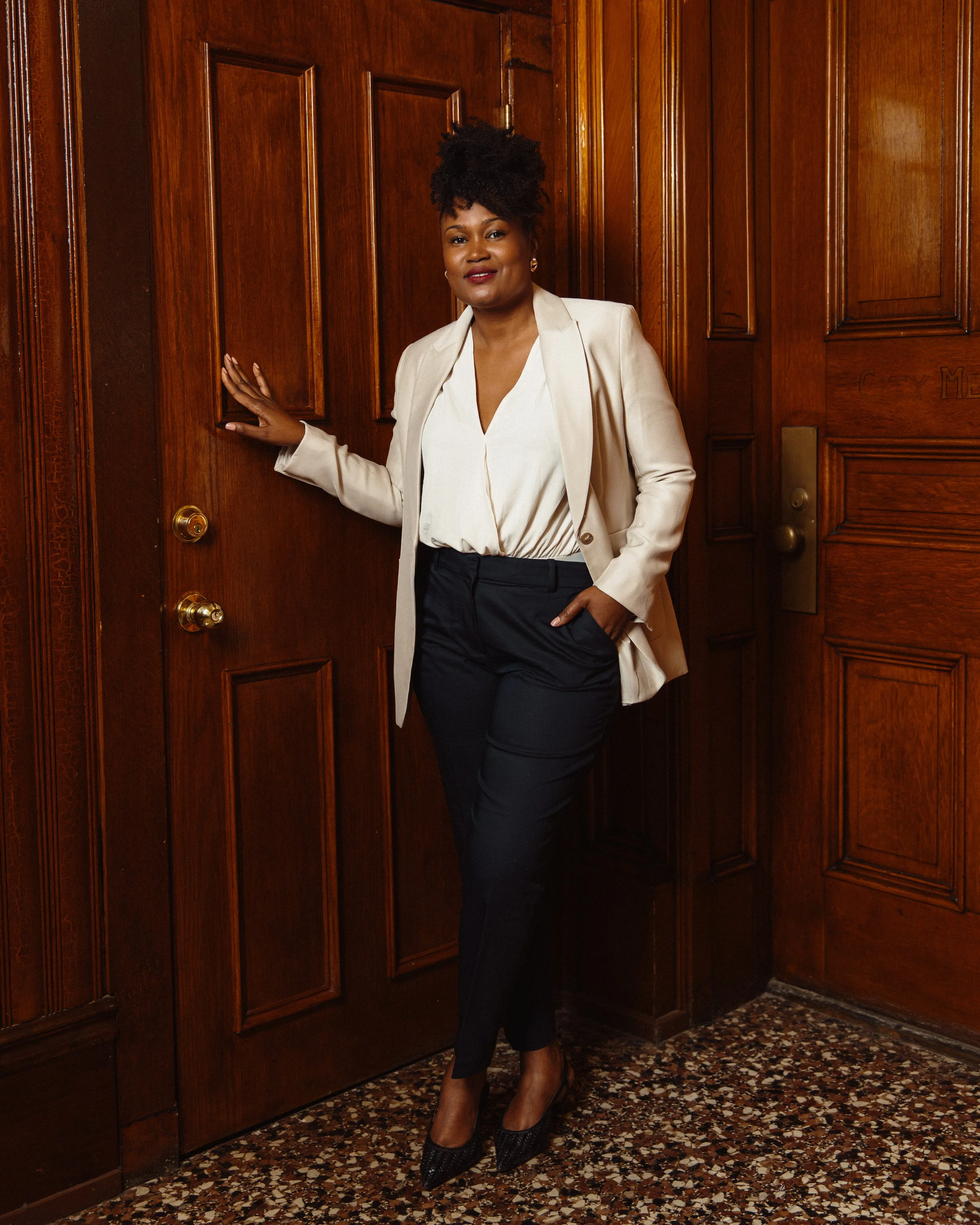 Dr.Ellie Paris Miranda in a white blazer, cream blouse, and black pants standing against a wooden-paneled wall, with her hand on the door.