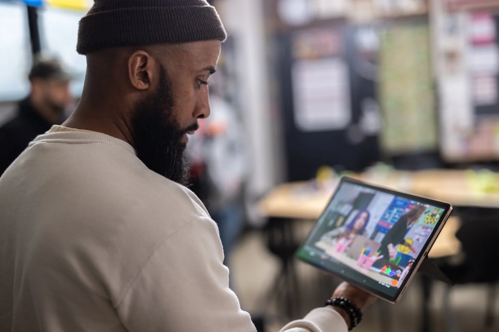 A man with a beard wearing a beanie and a white shirt, holding a tablet and looking at colorful images on the screen, seated in a room with large windows and blurred background.