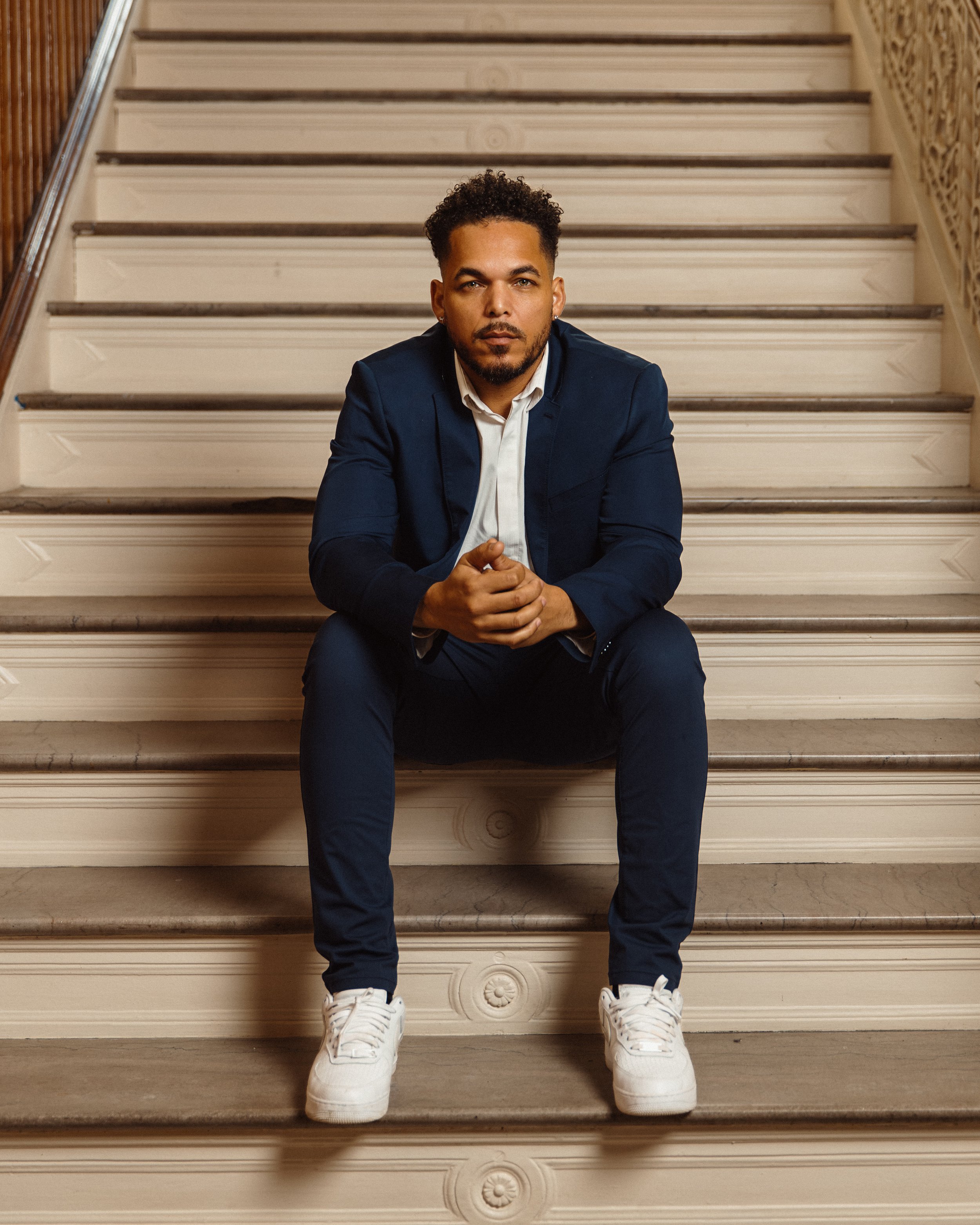 Salo Afonseca with curly hair, beard, and earrings sitting on stairs, wearing a dark blue suit, white shirt, white sneakers, looking at camera.