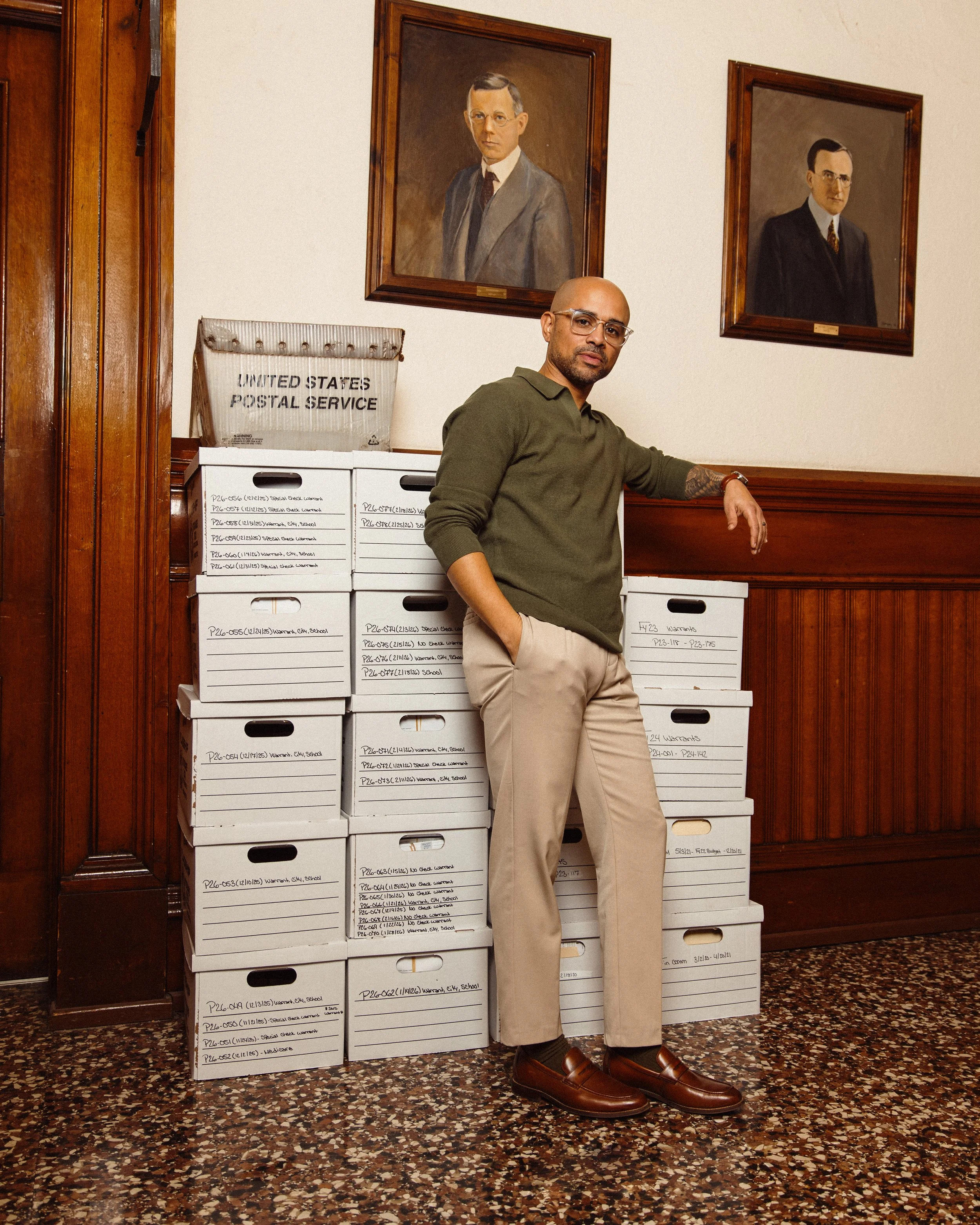 Nelson Fernandes, Deputy Chief of Staff with glasses, a beard, and tattoos on his arm leaning against a wall with a stack of white boxes labeled with various text, in a wood-paneled room with two portraits hanging on the wall behind him.