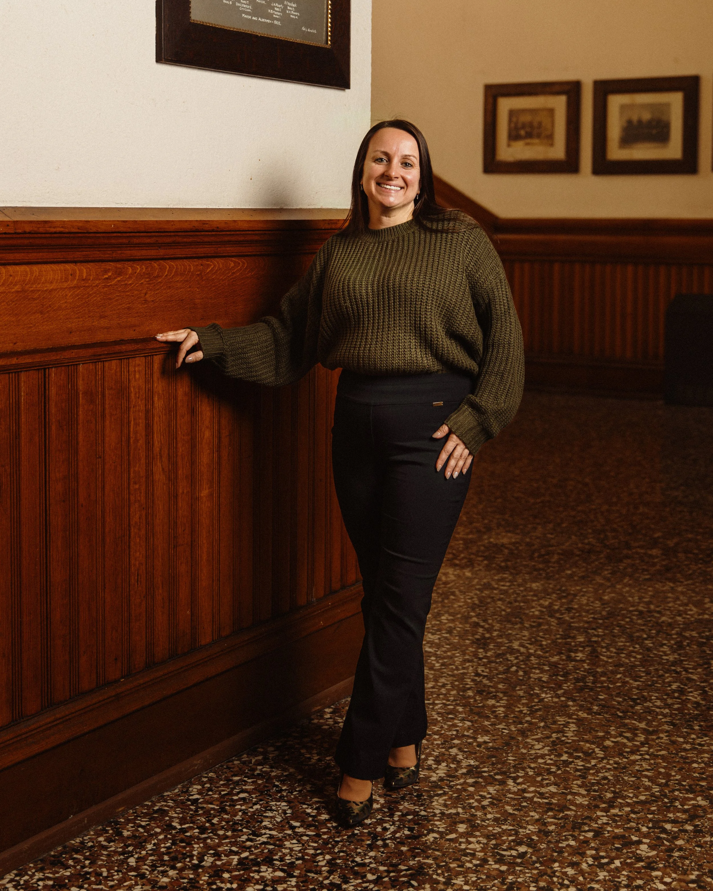 A woman with long brown hair wearing a green sweater and black pants standing next to a wooden-paneled wall inside a building with framed pictures on the wall and a patterned carpet on the floor.