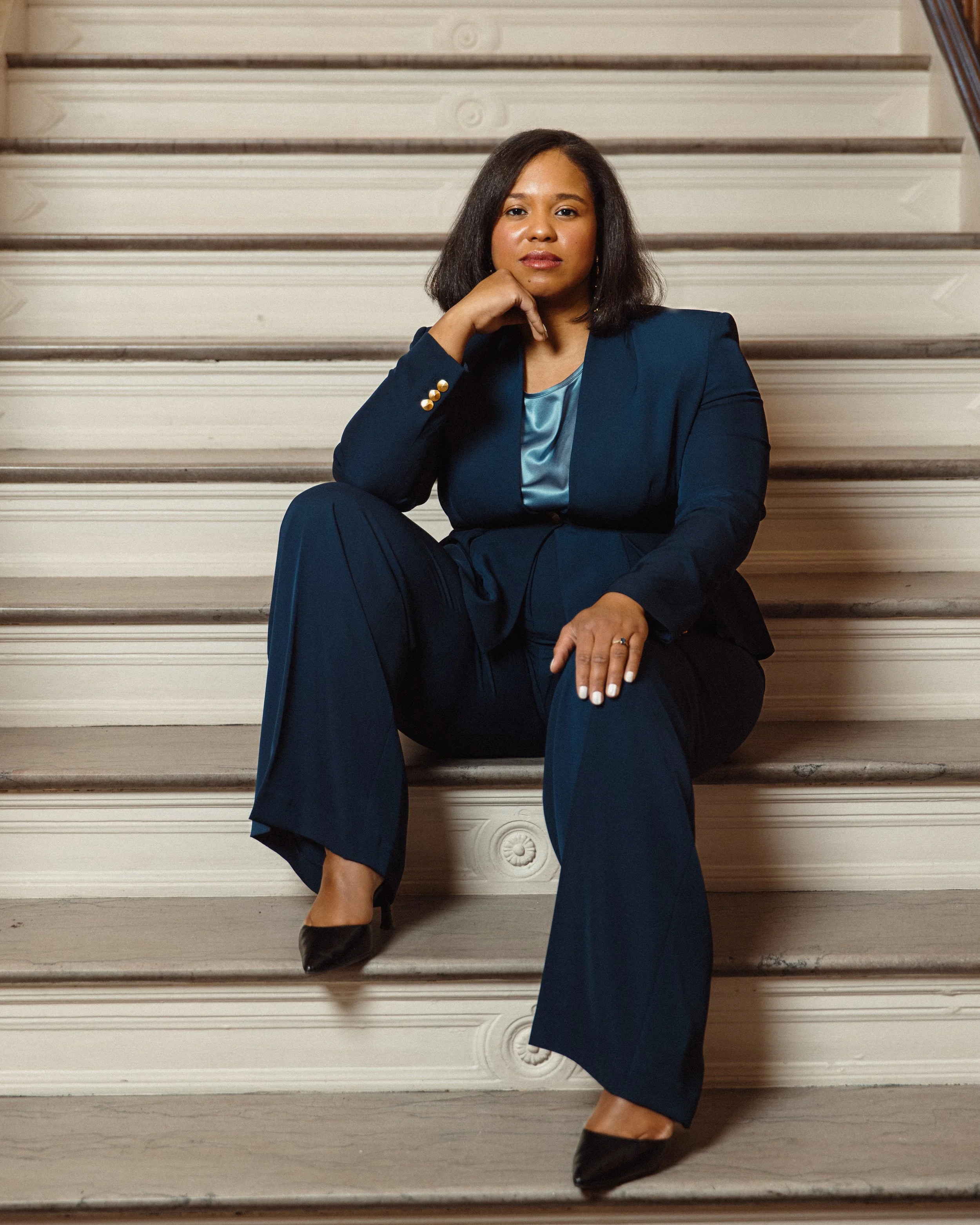 A woman sitting on stairs, wearing a dark blue suit and black shoes, with her hand posed under her chin.
