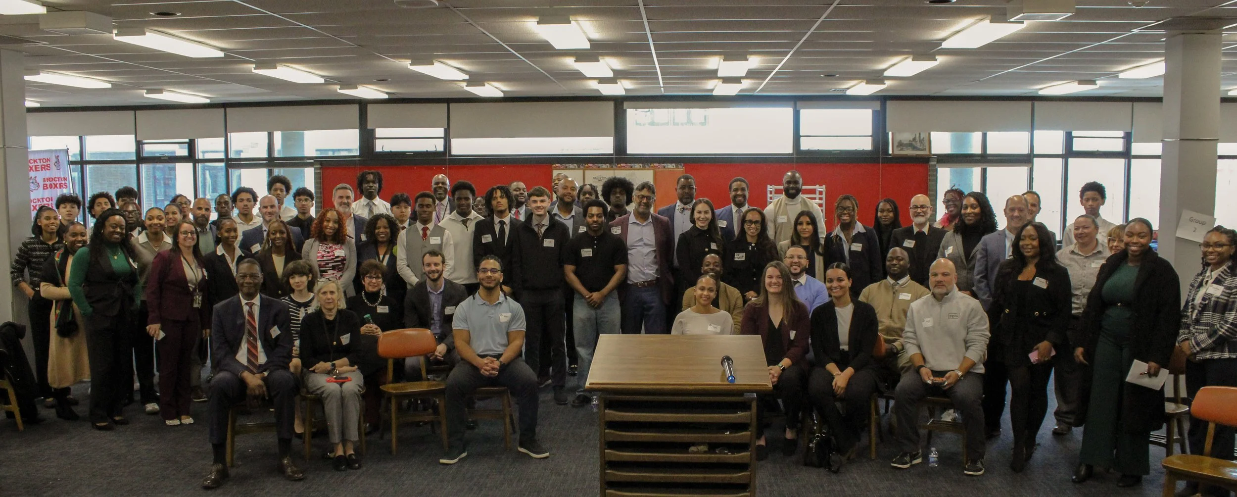 Group photo of diverse people gathered indoors for an event or meeting, with some seated and others standing, in a room with large windows and indoor lighting.