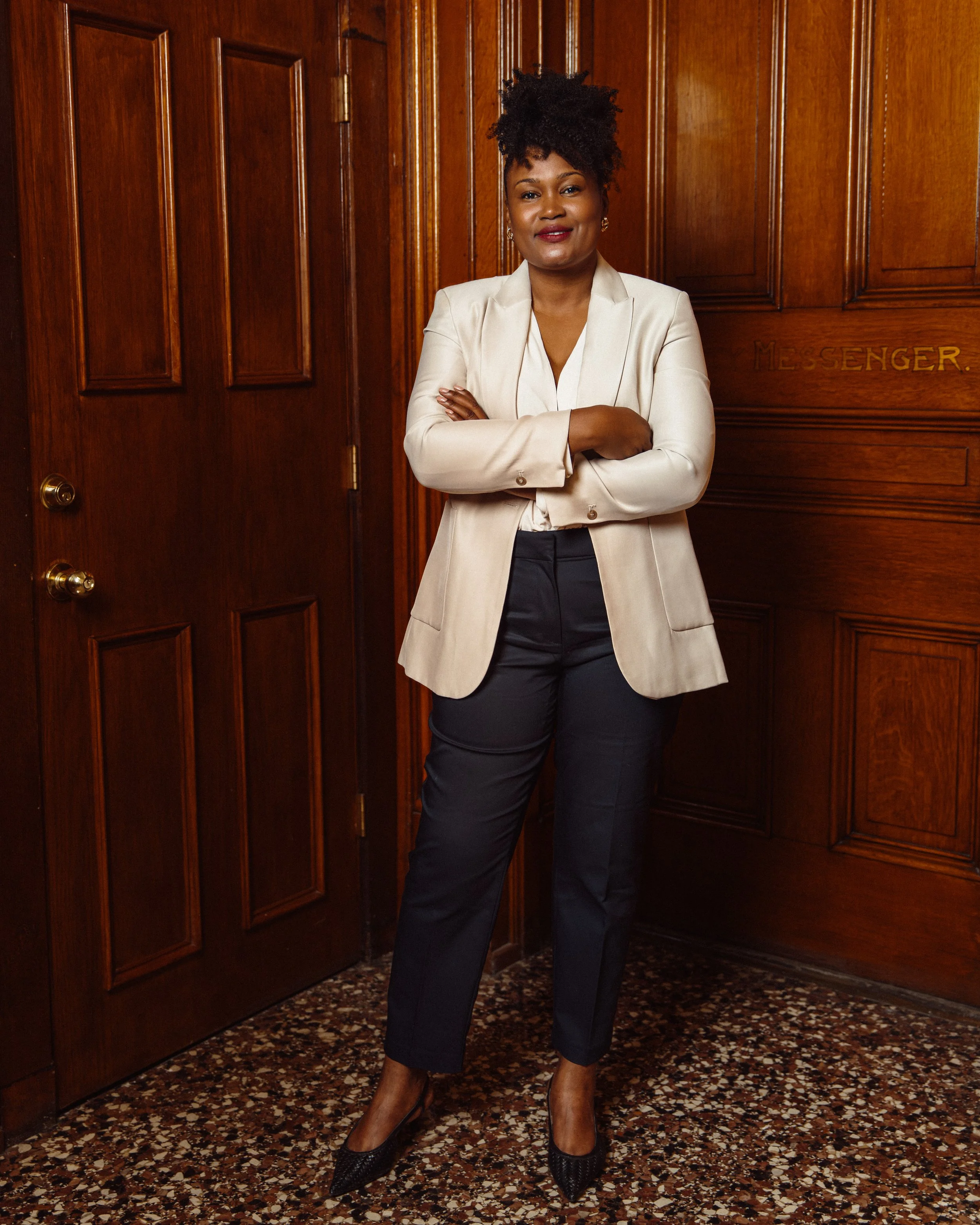 Dr.Ellie Paris Miranda  with dark skin and curly hair styled in an updo, standing with her arms crossed, dressed in a cream-colored blazer and navy pants, in front of a wooden paneled wall with the word 'MESSENGER' faintly visible.