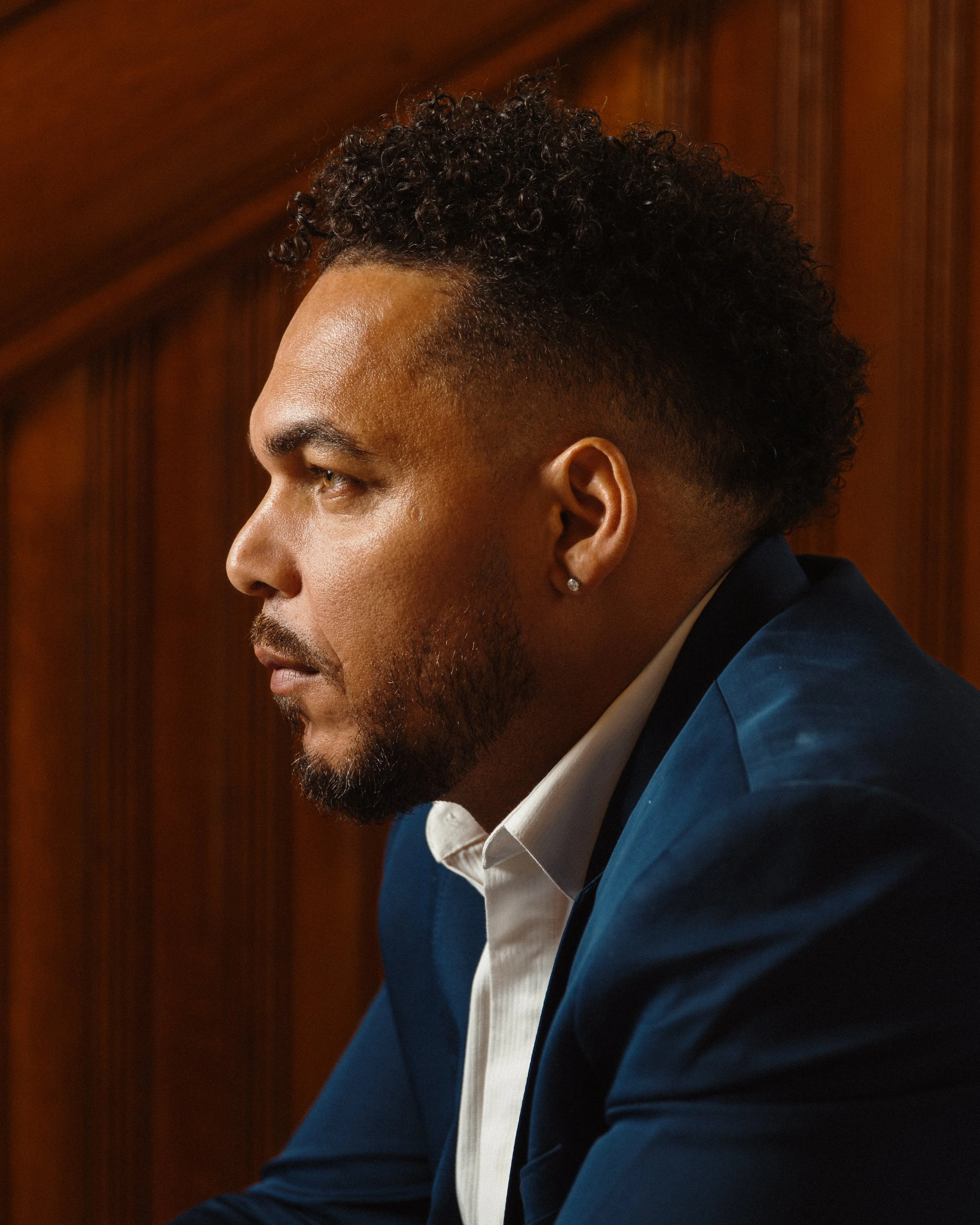 Close-up profile of Salo Afonseca with curly hair, wearing a blue blazer, white shirt, and earring, sitting in front of a wooden background.