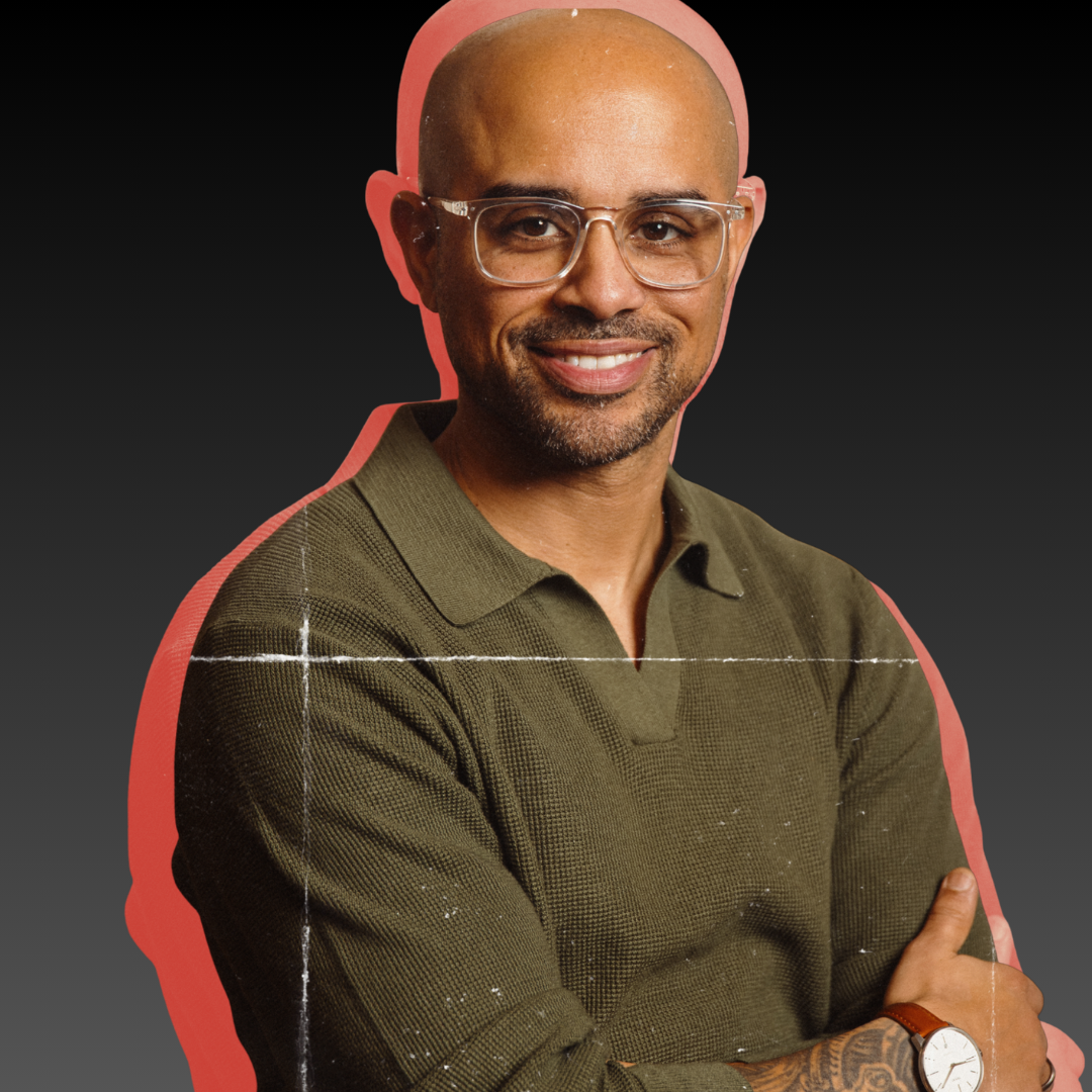 Nelson Fernandes with glasses, a bald head, wearing an olive green shirt, crossed arms, and a wristwatch, standing against a dark background with a pink outline.